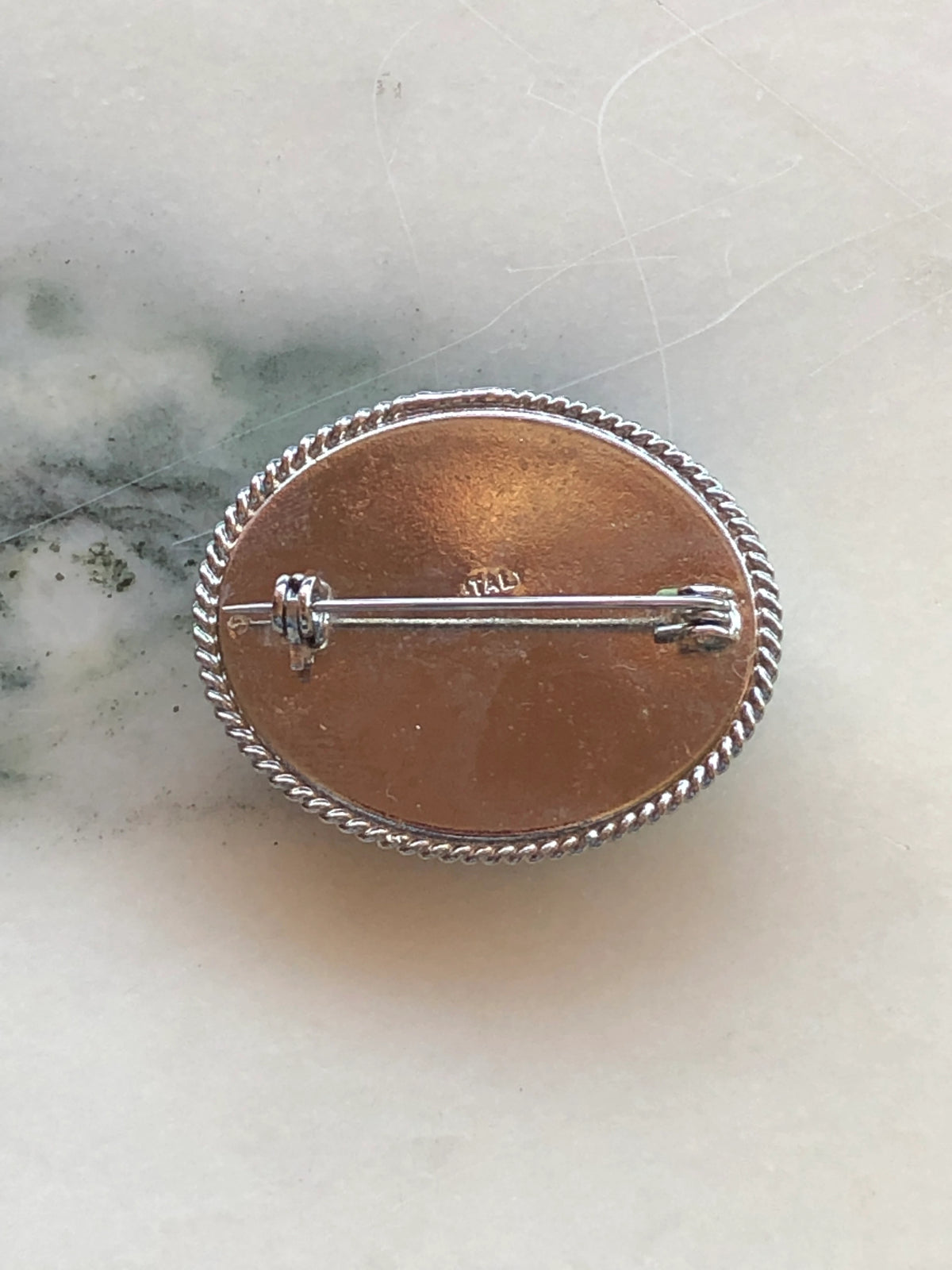 The back view of a A 3cm x 4cm vintage Italian micro mosaic oval brooch featuring a hand assembled glass tessera design of forget-me-not flowers in cornflower blue, white and yellow on a blue ground, set within a silver-tone frame with twisted rope edge detail, photographed on a white marble table