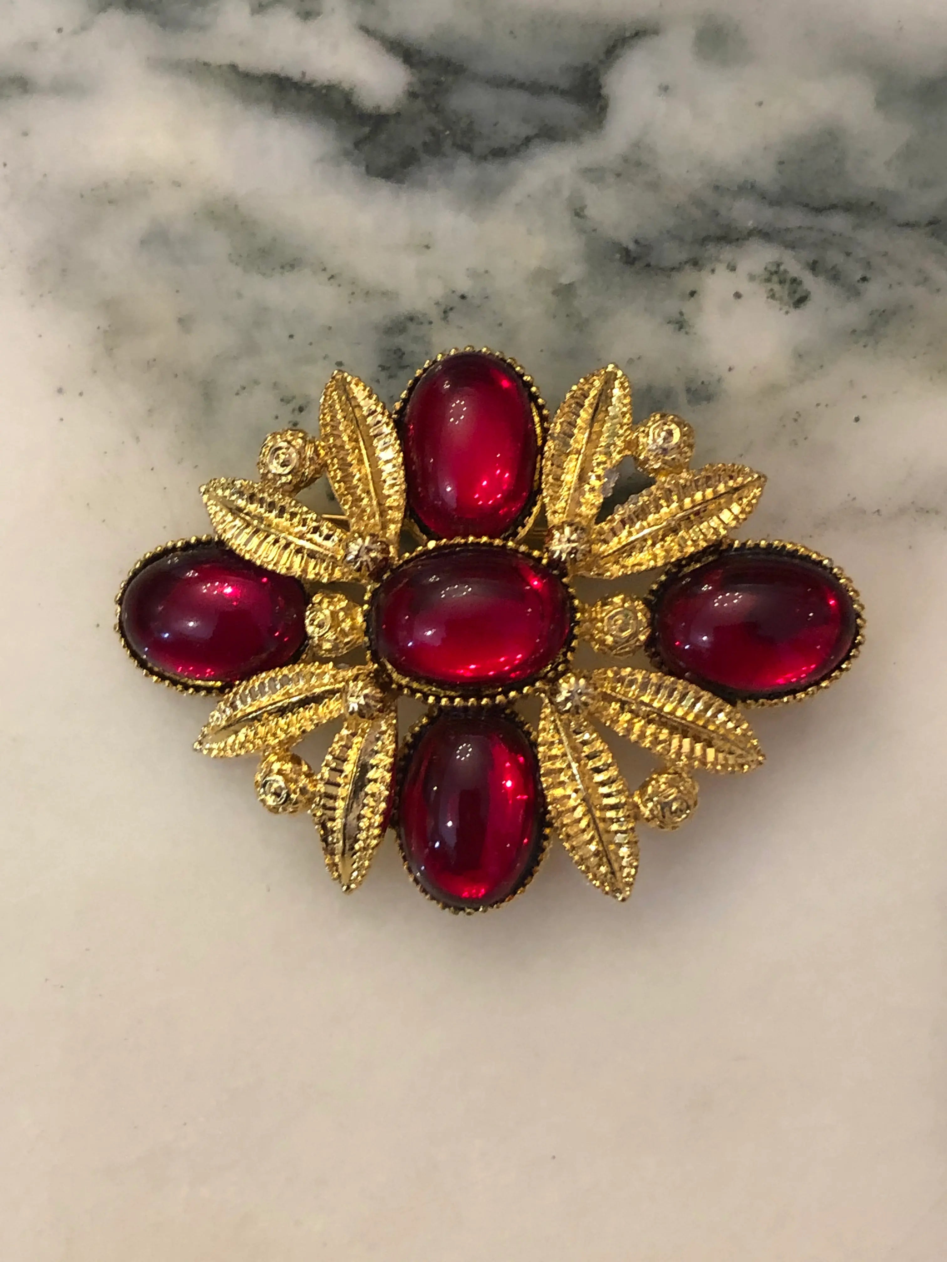 Broochella Fiorella Vintage Ruby Cabochon Brooch featuring five red oval cabochons surrounded by textured gold-tone leaves and scrollwork on a cream and grey marble background.