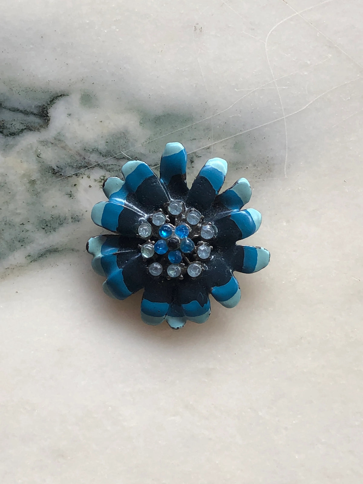 Broochella Fleur de Saphir Vintage Enamel Brooch featuring layered black petals with turquoise and pale blue enamel tips and blue and clear crystal details, photographed on a white marble background with soft grey-green veining.