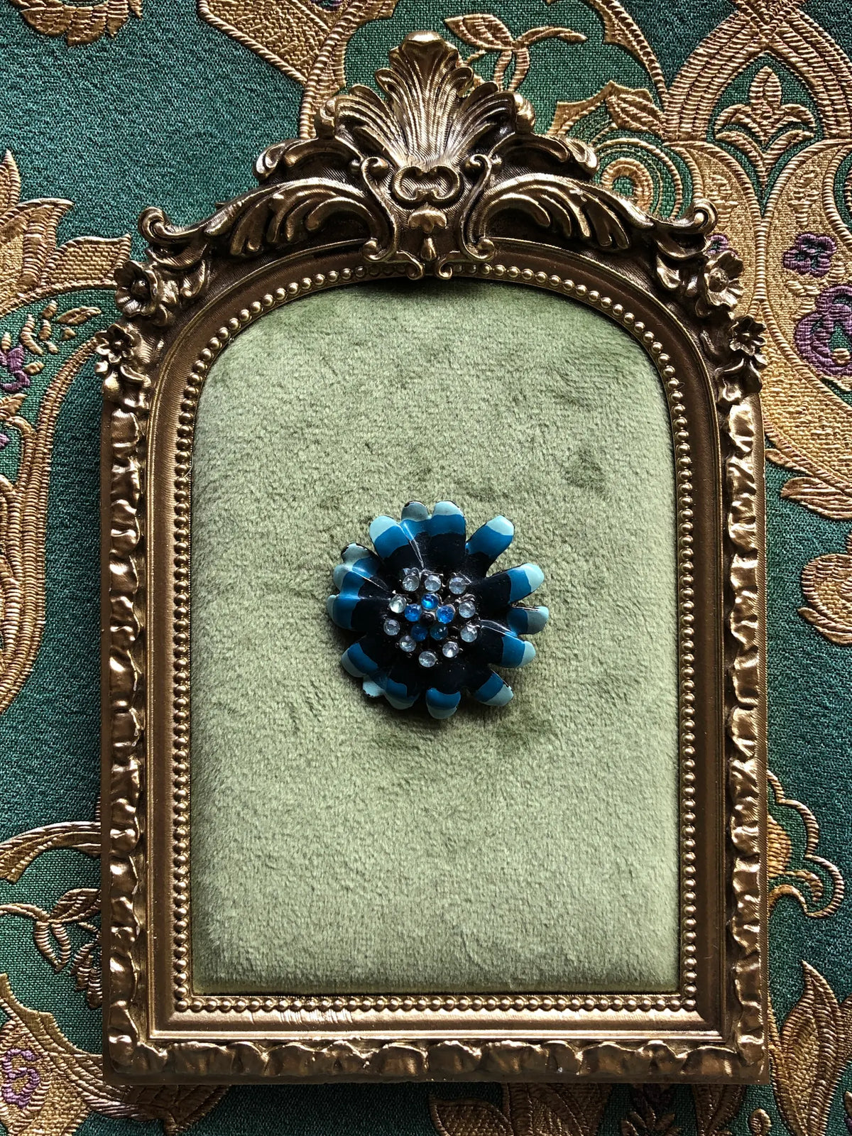 Broochella Fleur de Saphir Vintage Enamel Brooch featuring layered black petals tipped in vivid blue enamel with blue and clear crystal details, displayed on sage green velvet in an ornate antique gold frame against green and gold patterned fabric.