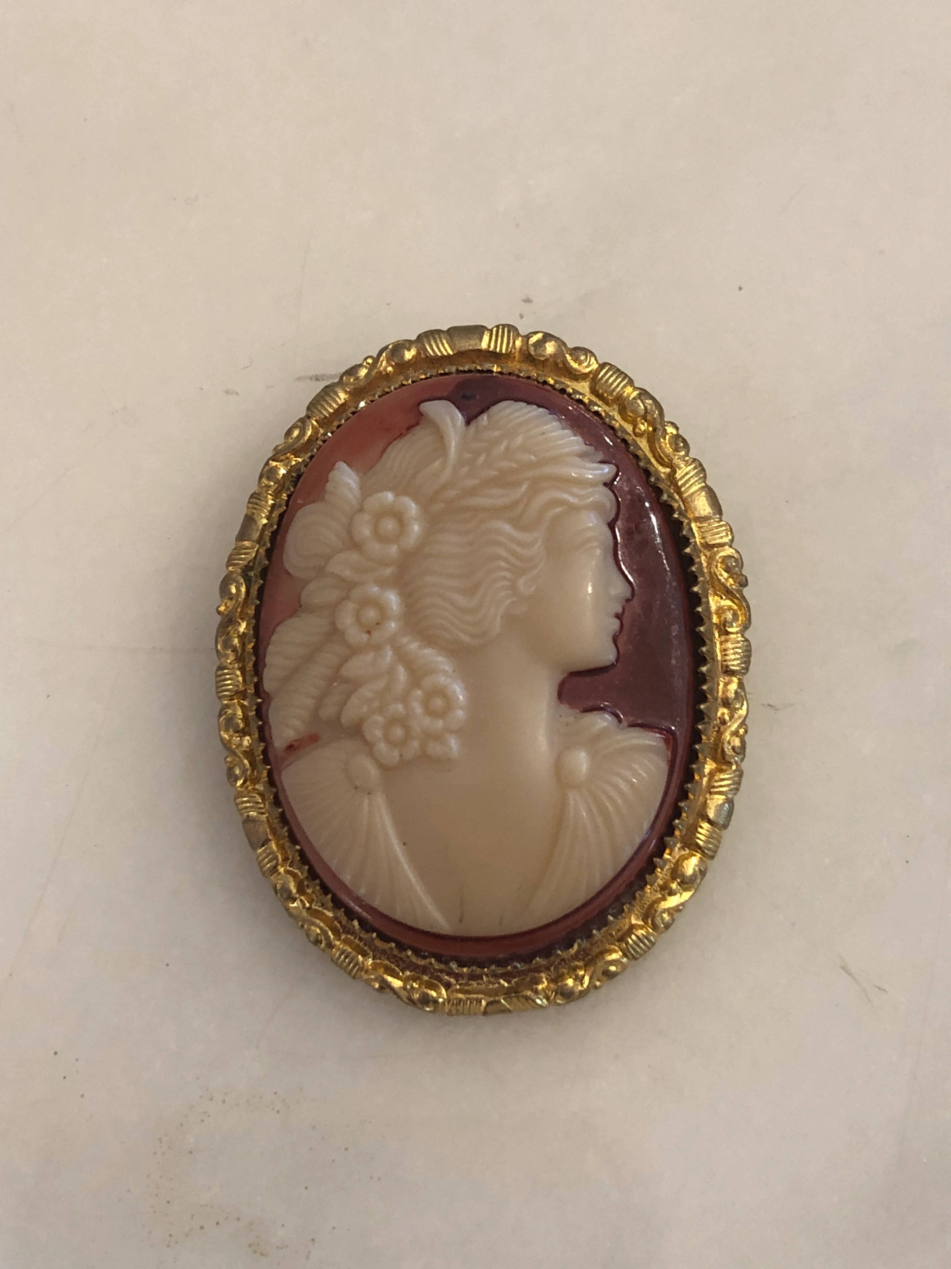 A Broochella Flora  4cm x 3cm vintage shell cameo brooch with a carved cream relief portrait of a flower-crowned goddess in profile on a pink-mauve ground, set in a gold-tone scrollwork frame. The background is white marble