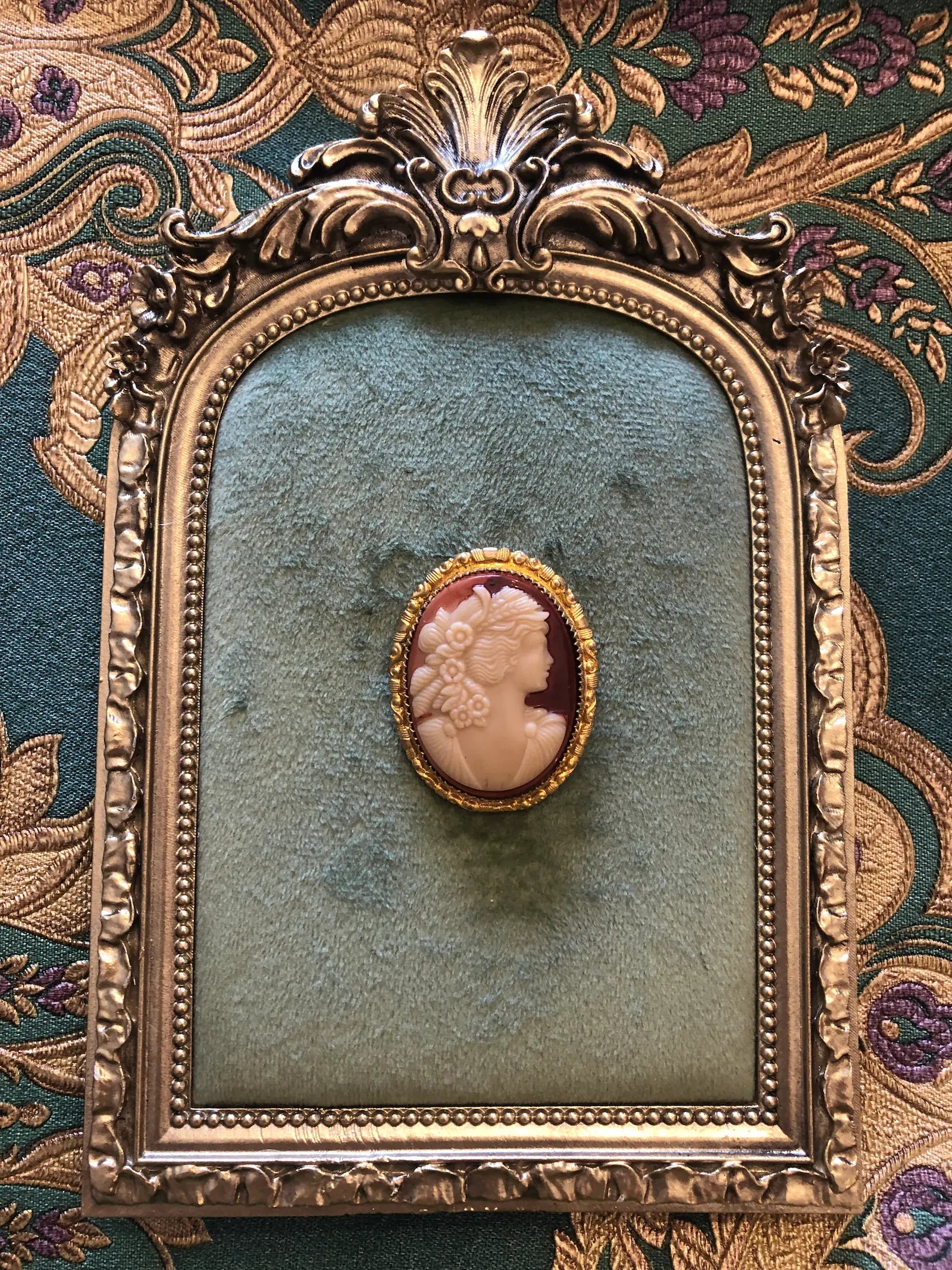 A Broochella Flora  4cm x 3cm vintage shell cameo brooch with a carved cream relief portrait of a flower-crowned goddess in profile on a pink-mauve ground, set in a gold-tone scrollwork frame. Photographed on a green velvet tray with an ornate gold frame. The background is green and gold damask wallpaper