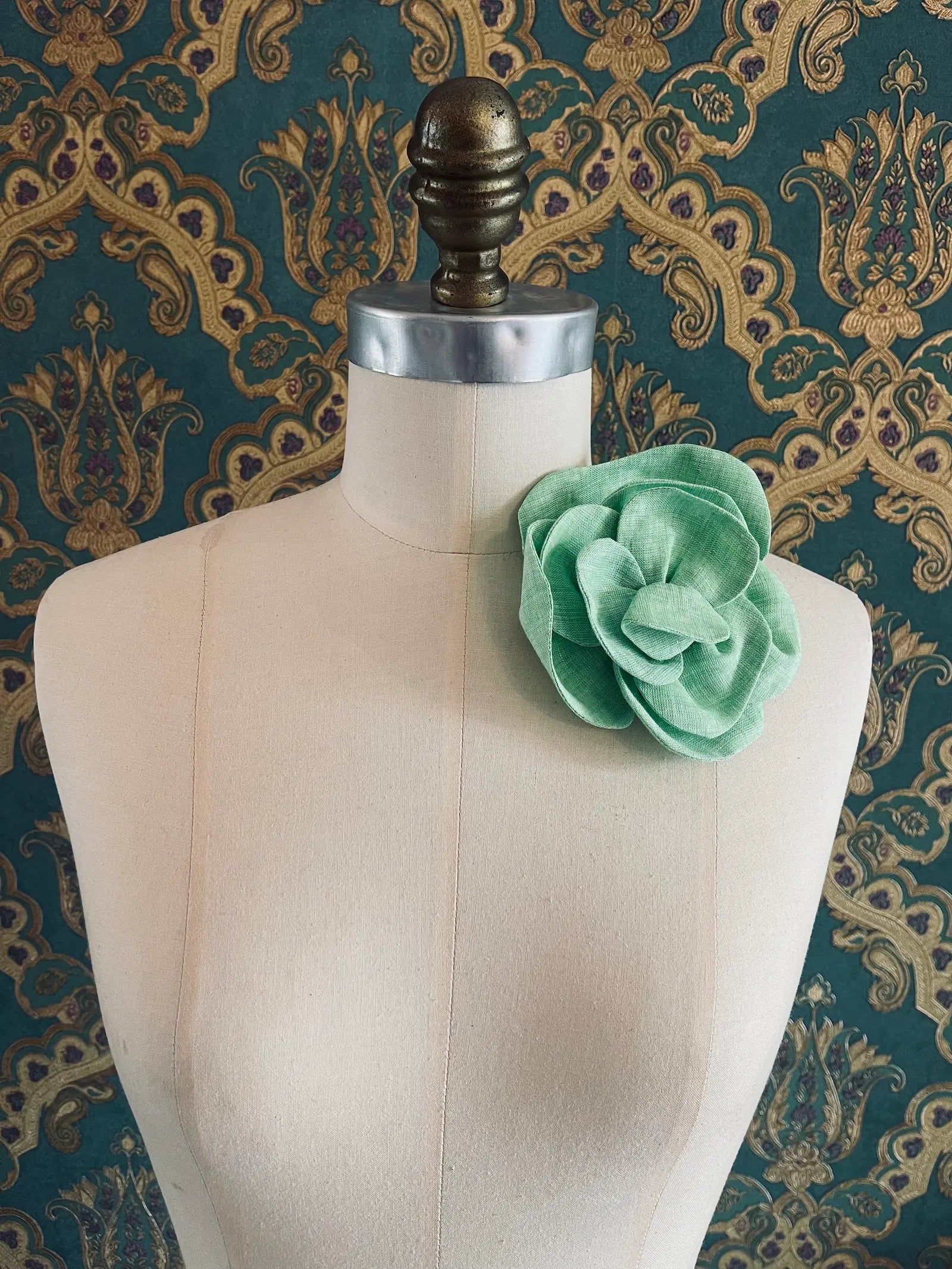 Broochella Florella small handmade linen flower brooch in green. Pictured on a mannequin. The background is green and gold damask wallpaper.
