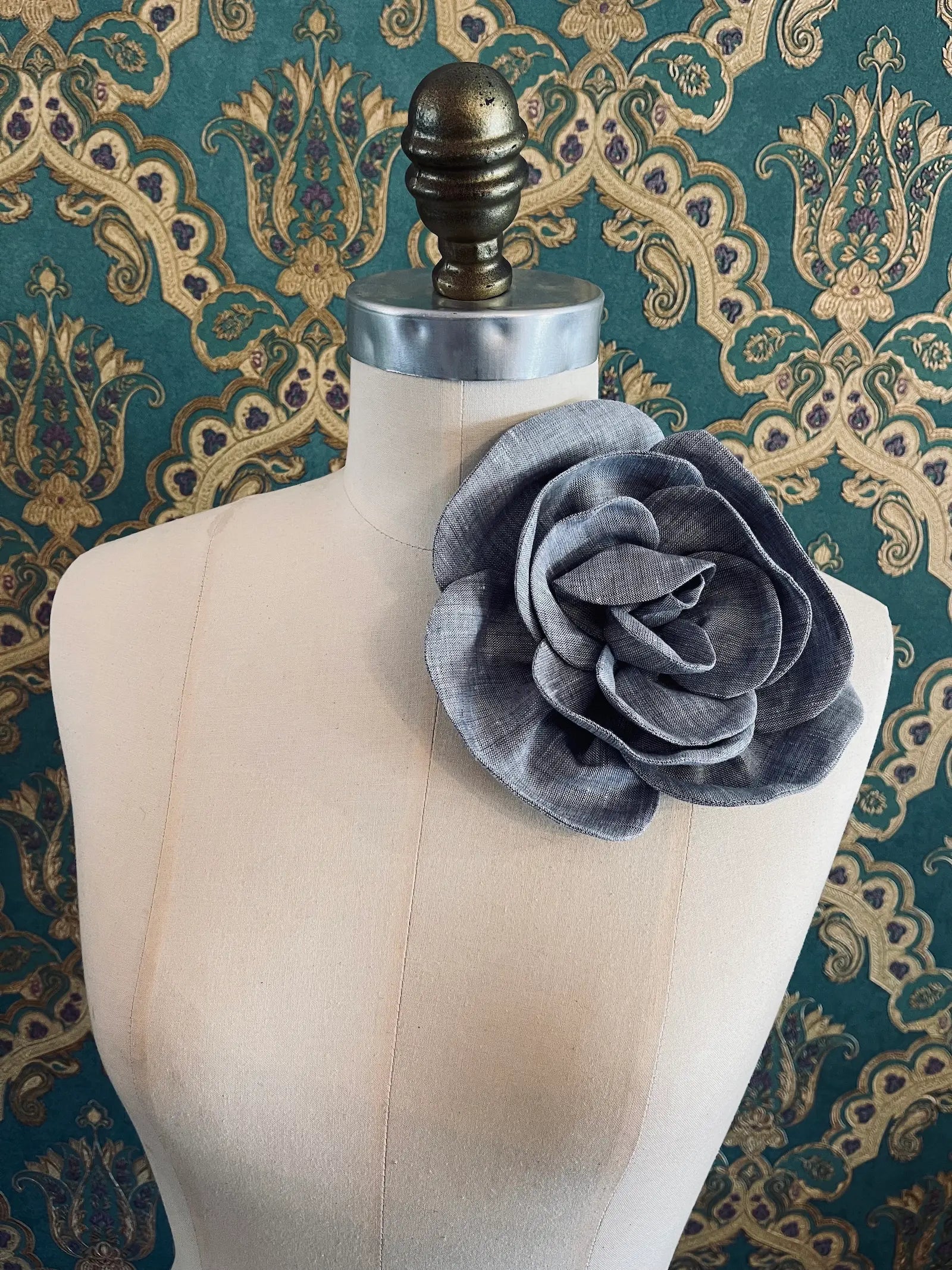Broochella Florella large handmade linen flower brooch in grey. Pictured on a mannequin. The background is green and gold damask wallpaper.