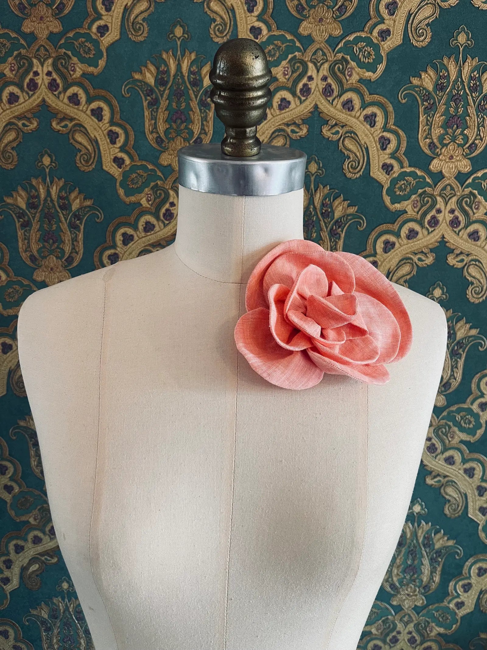 Broochella Florella small handmade linen flower brooch in orange. Pictured on a mannequin. The background is green and gold damask wallpaper.