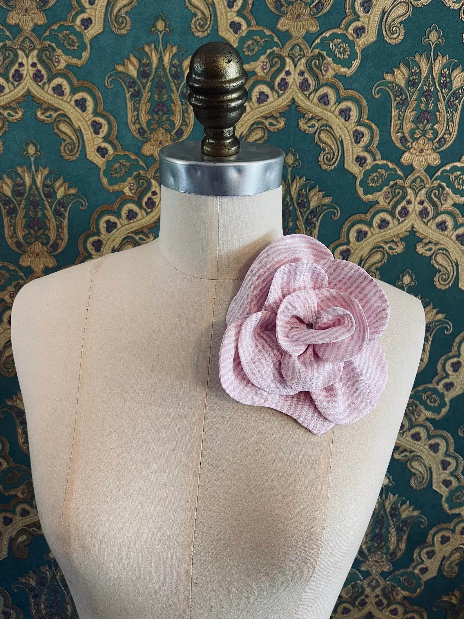 Broochella Florella pink handmade striped linen flower brooch. Pictured on a mannequin. The background is green and gold damask wallpaper