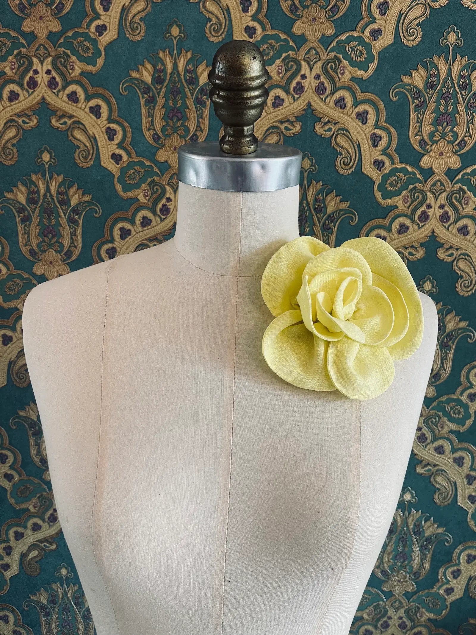 Broochella Florella small handmade linen flower brooch in yellow. Pictured on a mannequin. The background is green and gold damask wallpaper.