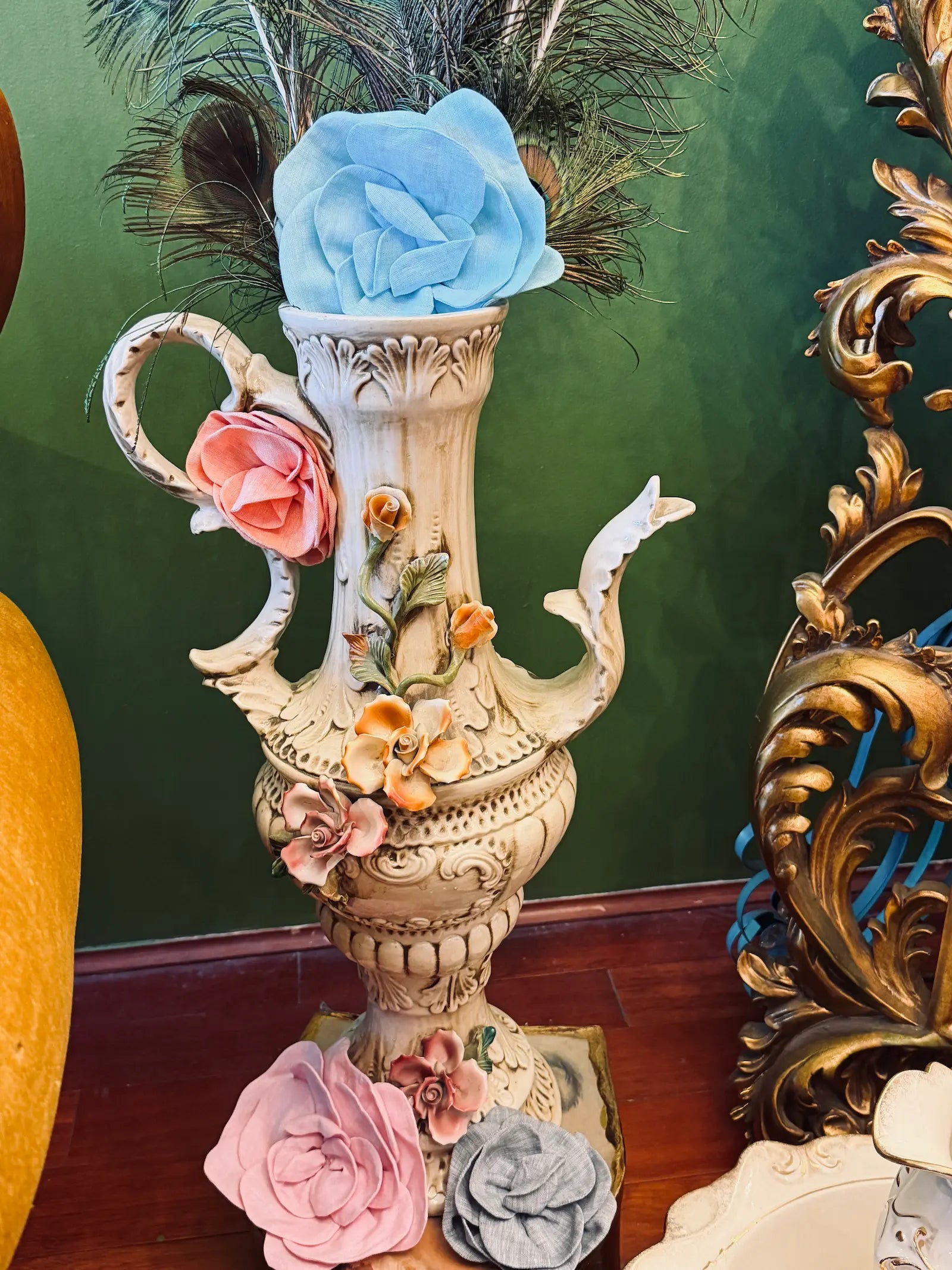 An ornate beige ceramic-style decorative vase with sculpted floral details, a handle, a spout, and faux peacock feathers inserted at the top, set against a dark green wall and wood floor. Placed on the vase are the Broochella Florella linen flower brooches in blu, pink, grey and orange. 
