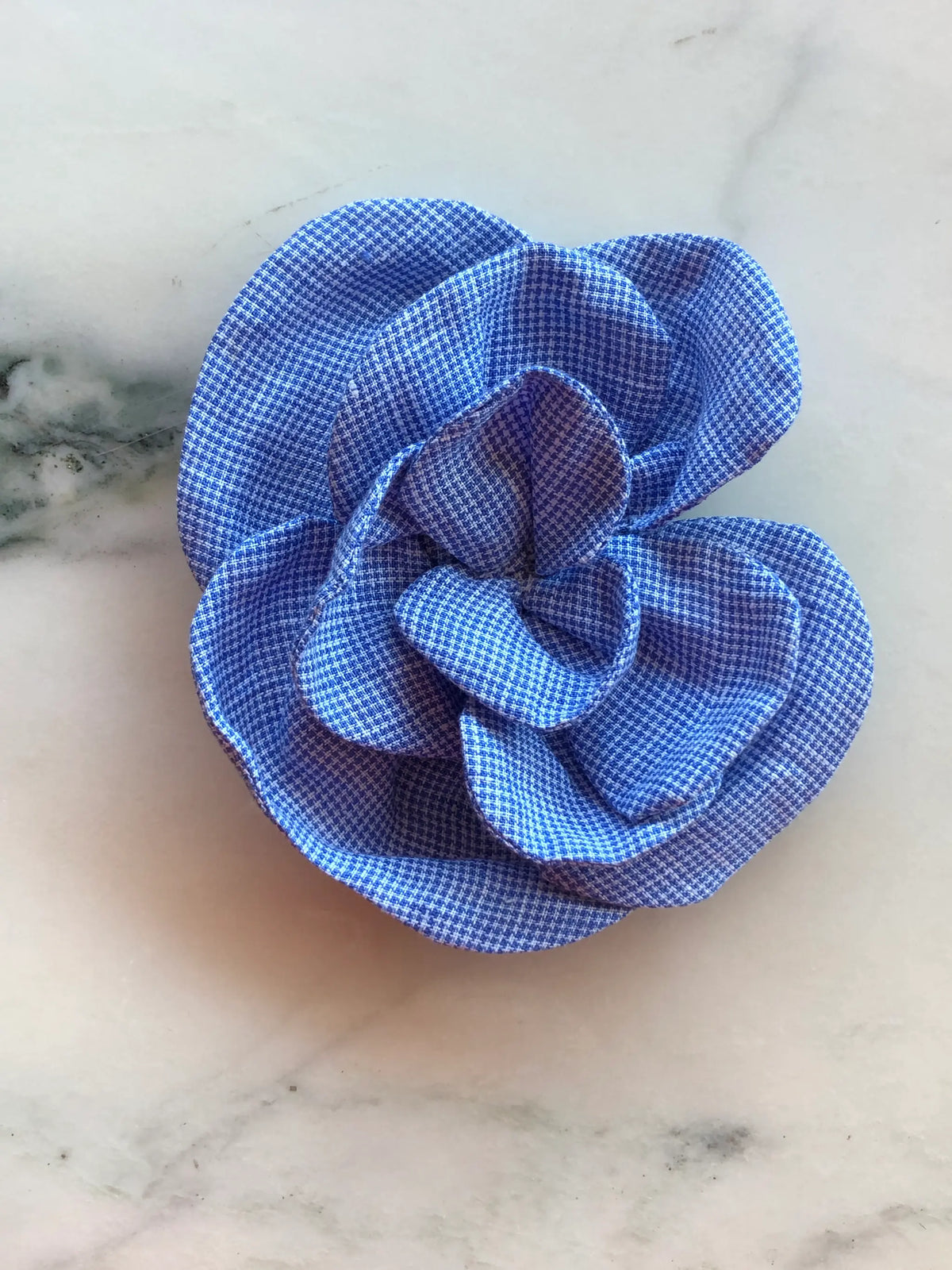 Broochella Florella blue and white checked linen flower brooch with sculptural layered petals, photographed on a white marble background with subtle grey veining.