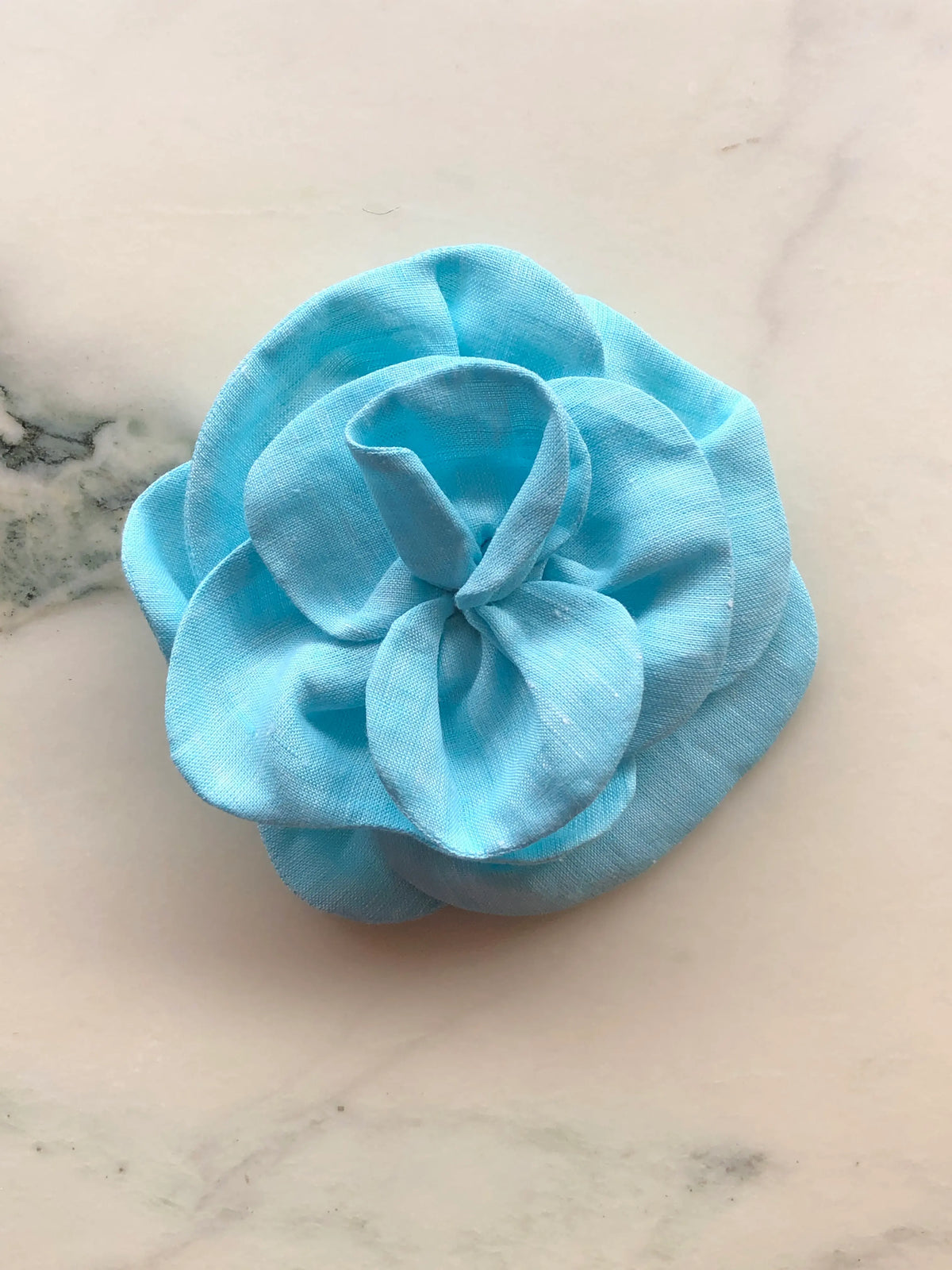 Small blue Broochella Florella Linen Flower Brooch with sculptural layered linen petals, photographed on a white marble background with soft grey veining.