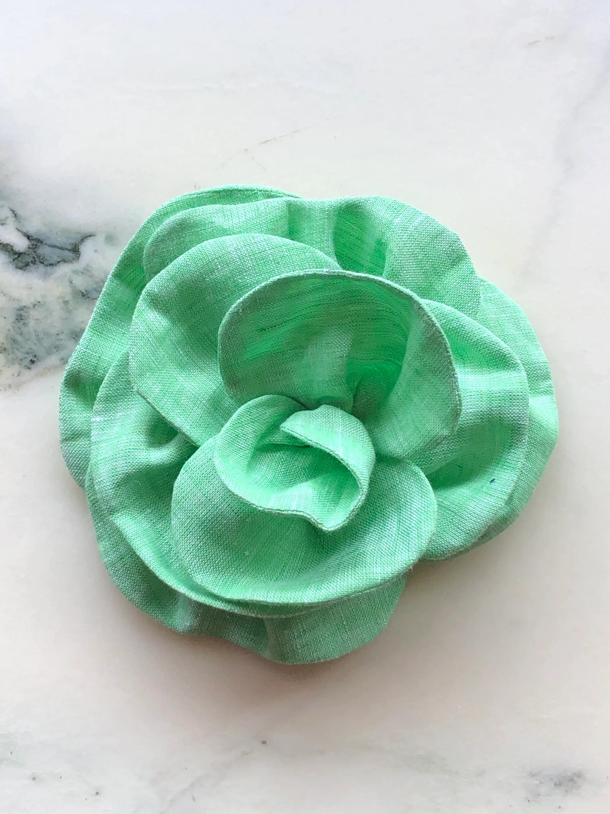 Small green Florella Linen Flower Brooch with sculptural layered linen petals, photographed on a white marble background with soft grey veining.
