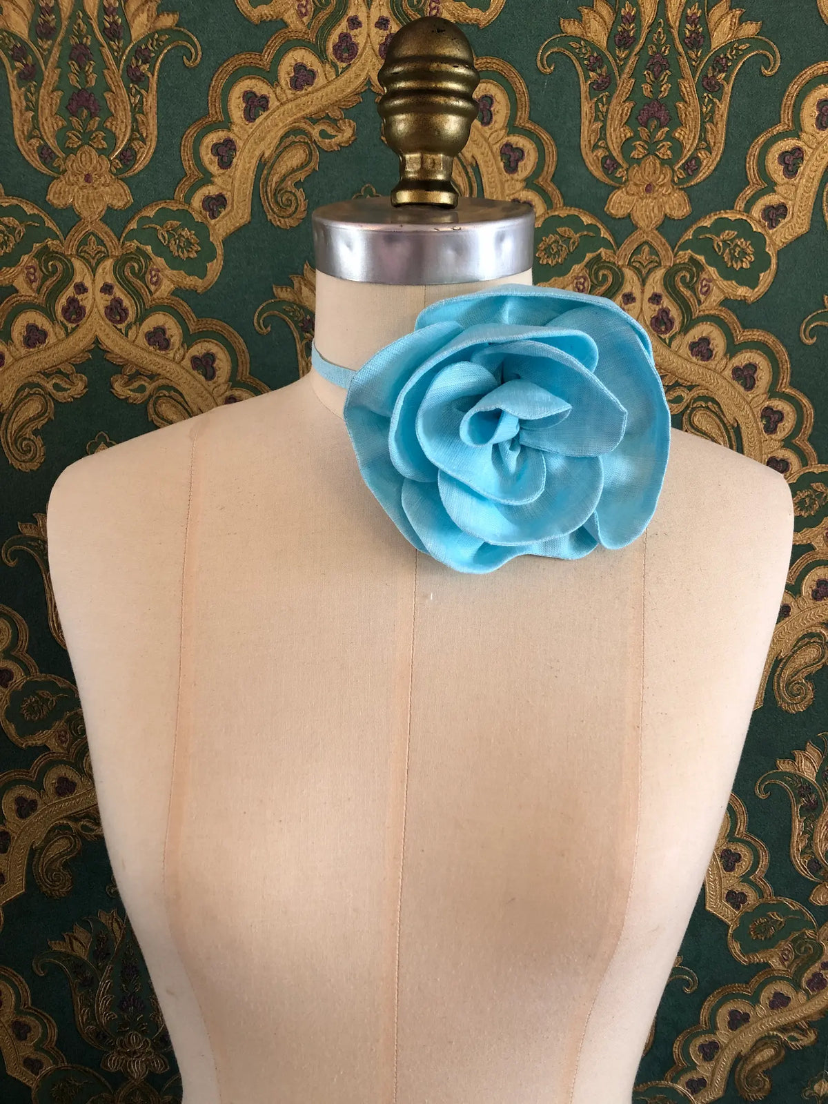 Broochella Florella small blue linen flower choker worn around the neck of a cream dress form against ornate teal and gold wallpaper.