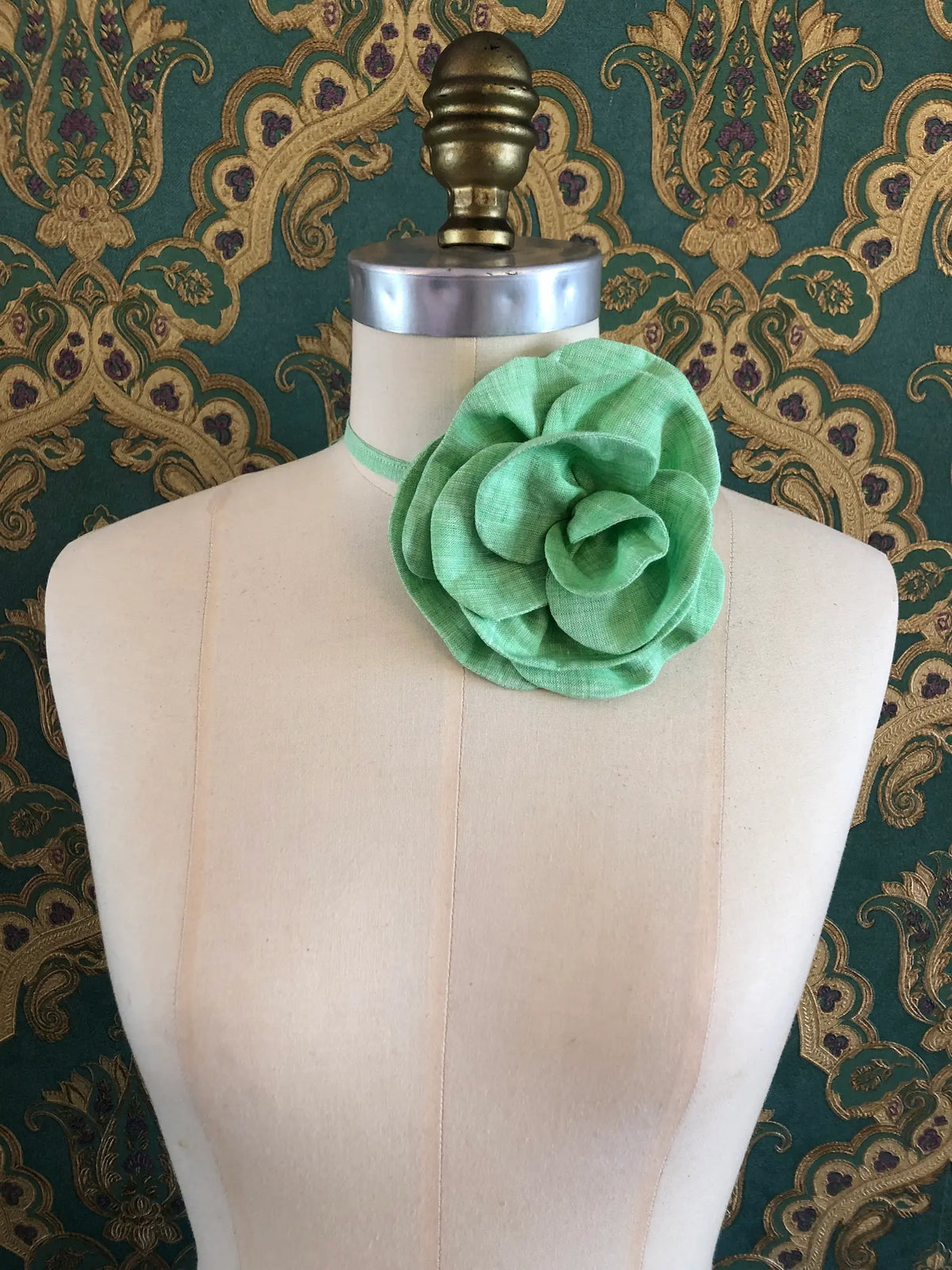 Broochella Florella small green linen flower choker worn around the neck of a cream dress form against ornate teal and gold wallpaper.