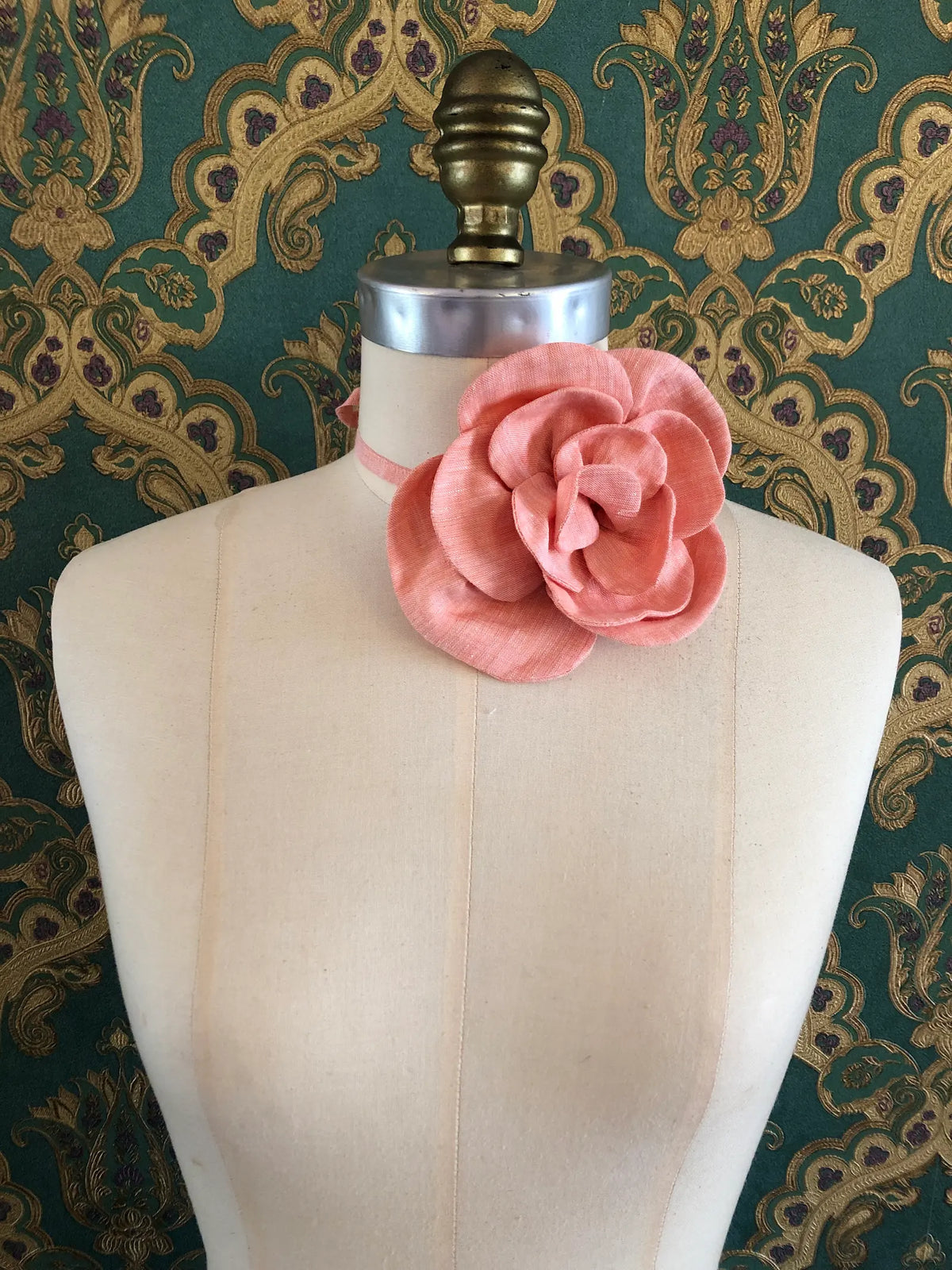 Broochella Florella small orange linen flower choker worn around the neck of a cream dress form against ornate teal and gold wallpaper.
