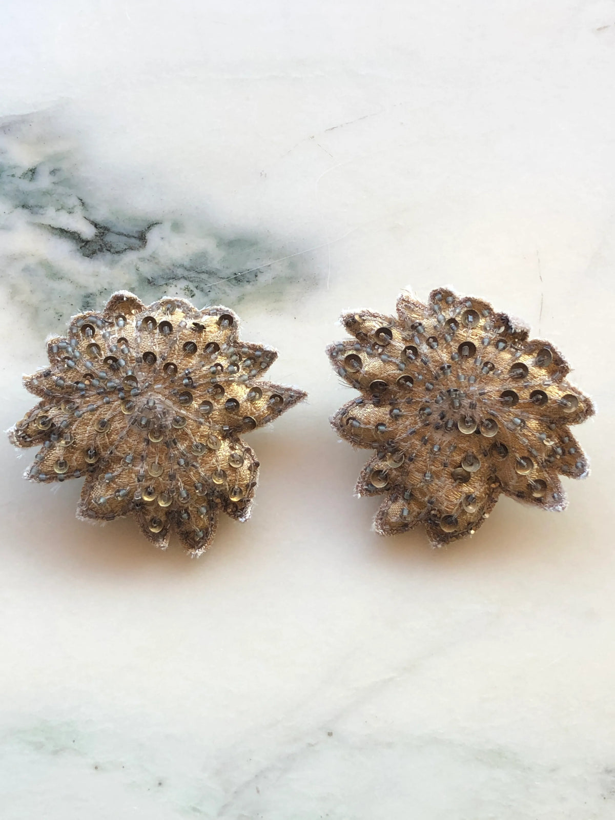 Broochella Foglie d'Autunno Leaf Brooch Pin set featuring two layered gold-toned embroidered brooches embellished with sequins and beads, photographed on a white marble background with soft grey veining.