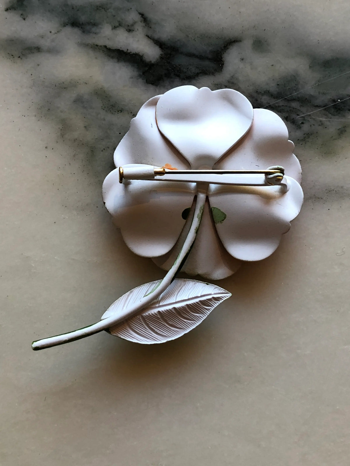 The back view of Broochella Francesca Vintage Orange Enamel Flower Brooch featuring layered orange enamel petals, black and white detailing and a green leaf and stem on a cream and grey marble background.