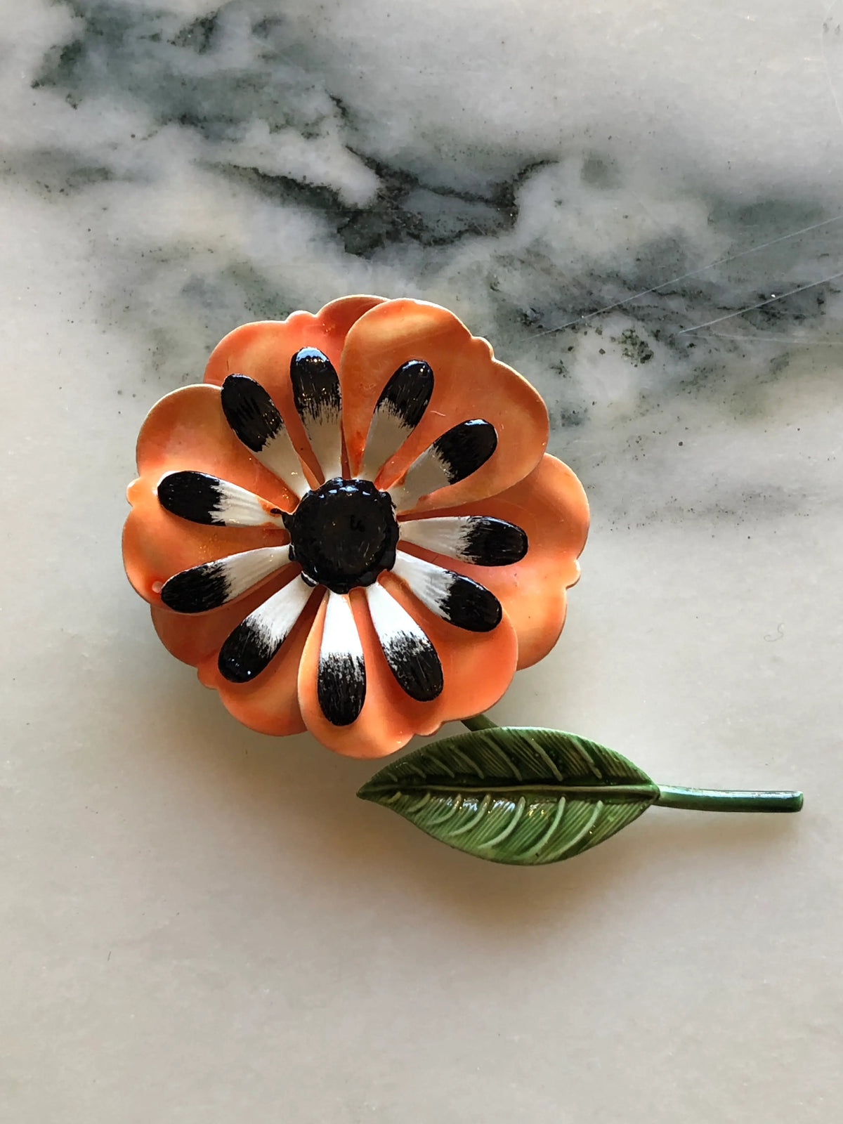 Broochella Francesca Vintage Orange Enamel Flower Brooch featuring layered orange enamel petals, black and white detailing and a green leaf and stem on a cream and grey marble background.