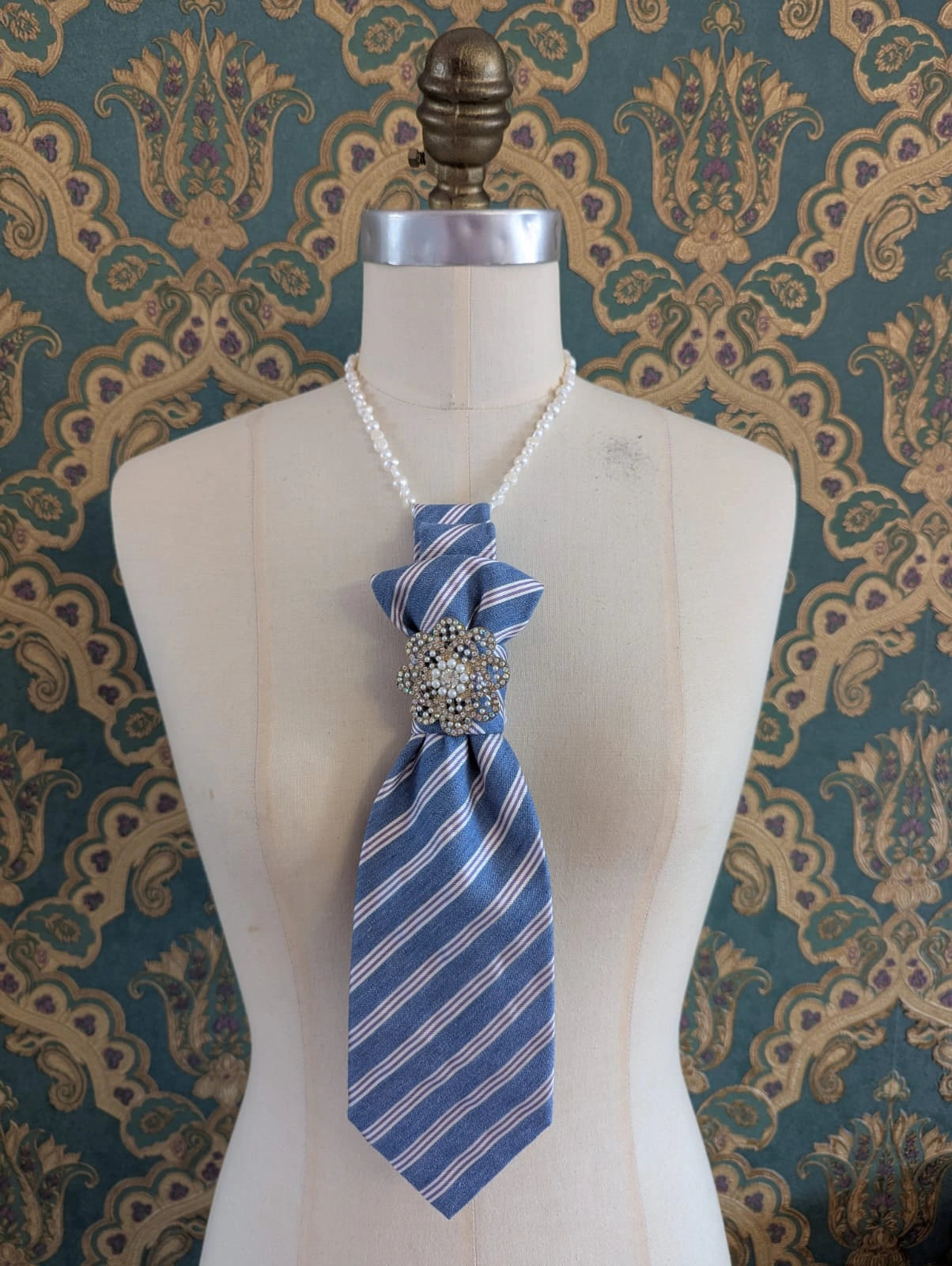 Broochella freshwater pearl 45 cm necklace. It is pictured on a mannequin, with the Broochella Montecristo upcycled tie brooch. The background is green and gold damask wallpaper.