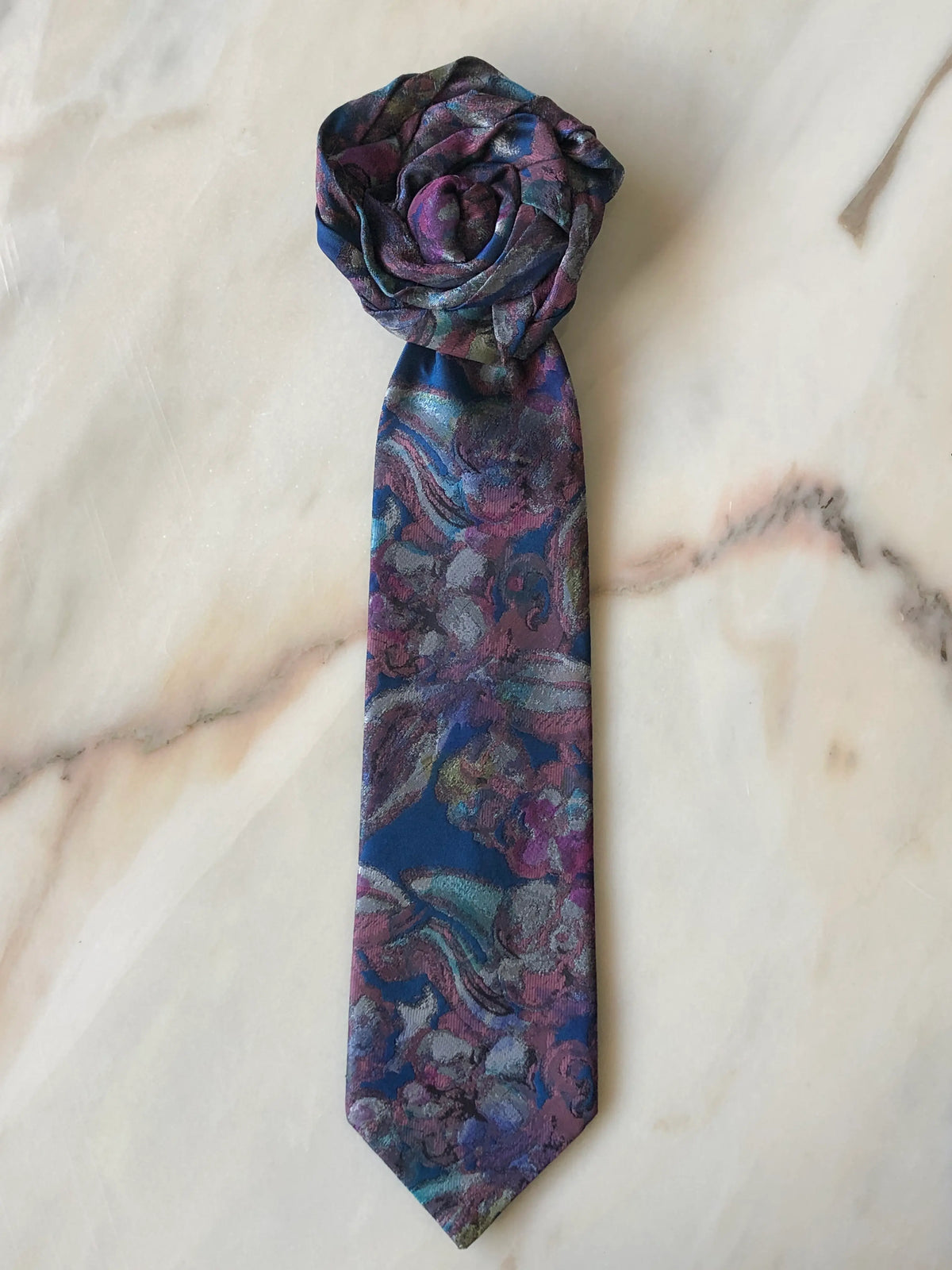 Broochella Fuente Rosette Tie Brooch - Saverio in a rich blue, plum and teal floral pattern, styled with a sculptural rosette and photographed on a white marble background.