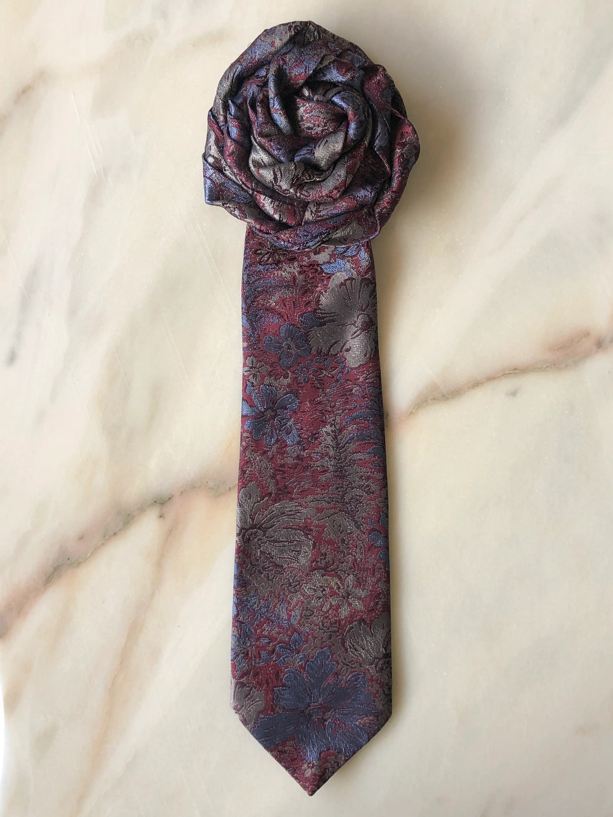 Broochella Fuente Rosette Tie Brooch - Stefano in burgundy, slate blue and grey floral jacquard, finished with a sculptural rosette and photographed on a white marble background.