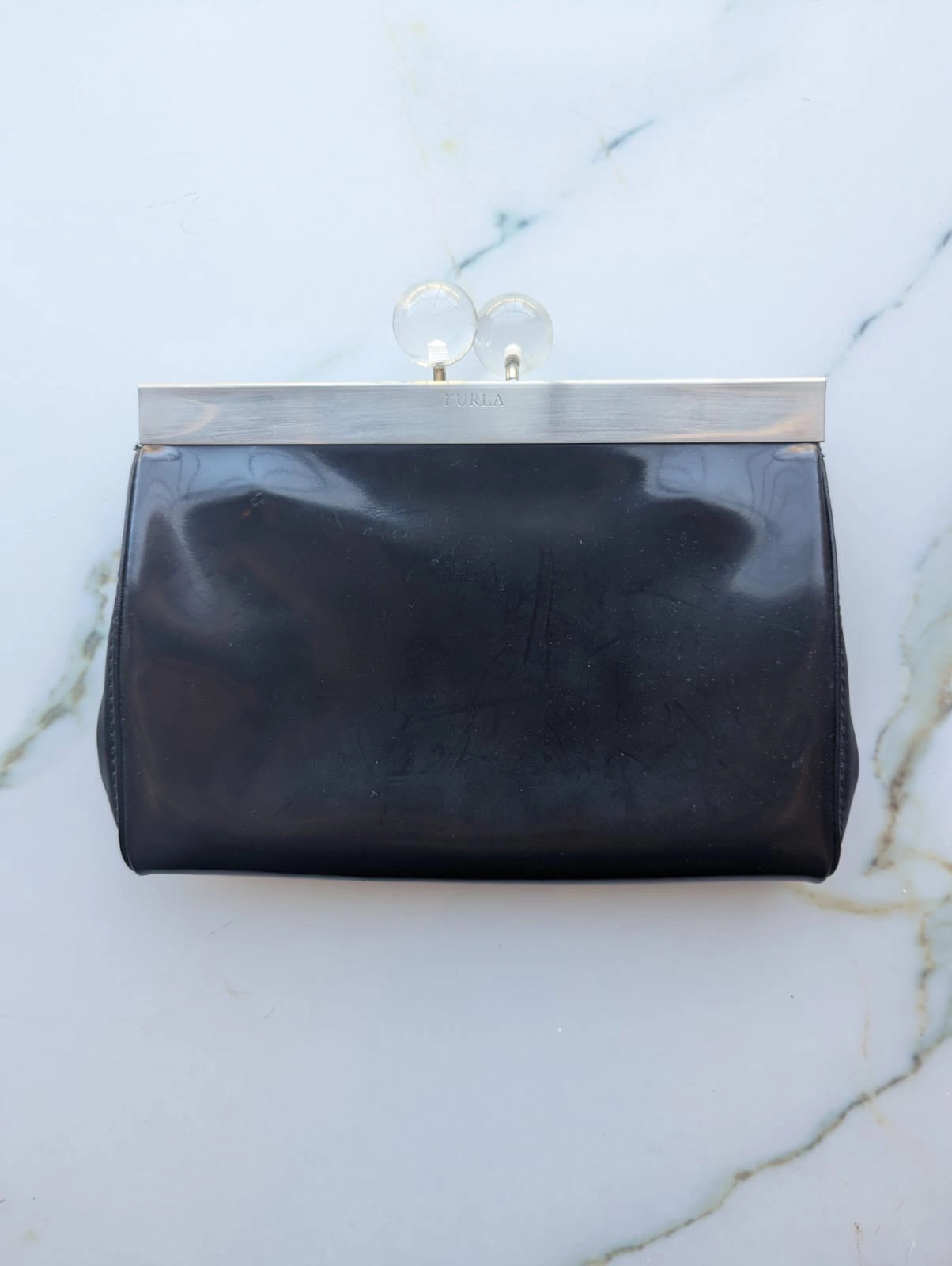 A Broochella The Stylist's Closet Furla black leather clutch purse with a silver-tone metal frame and clear acrylic clasp, displayed flat on a white marble surface.