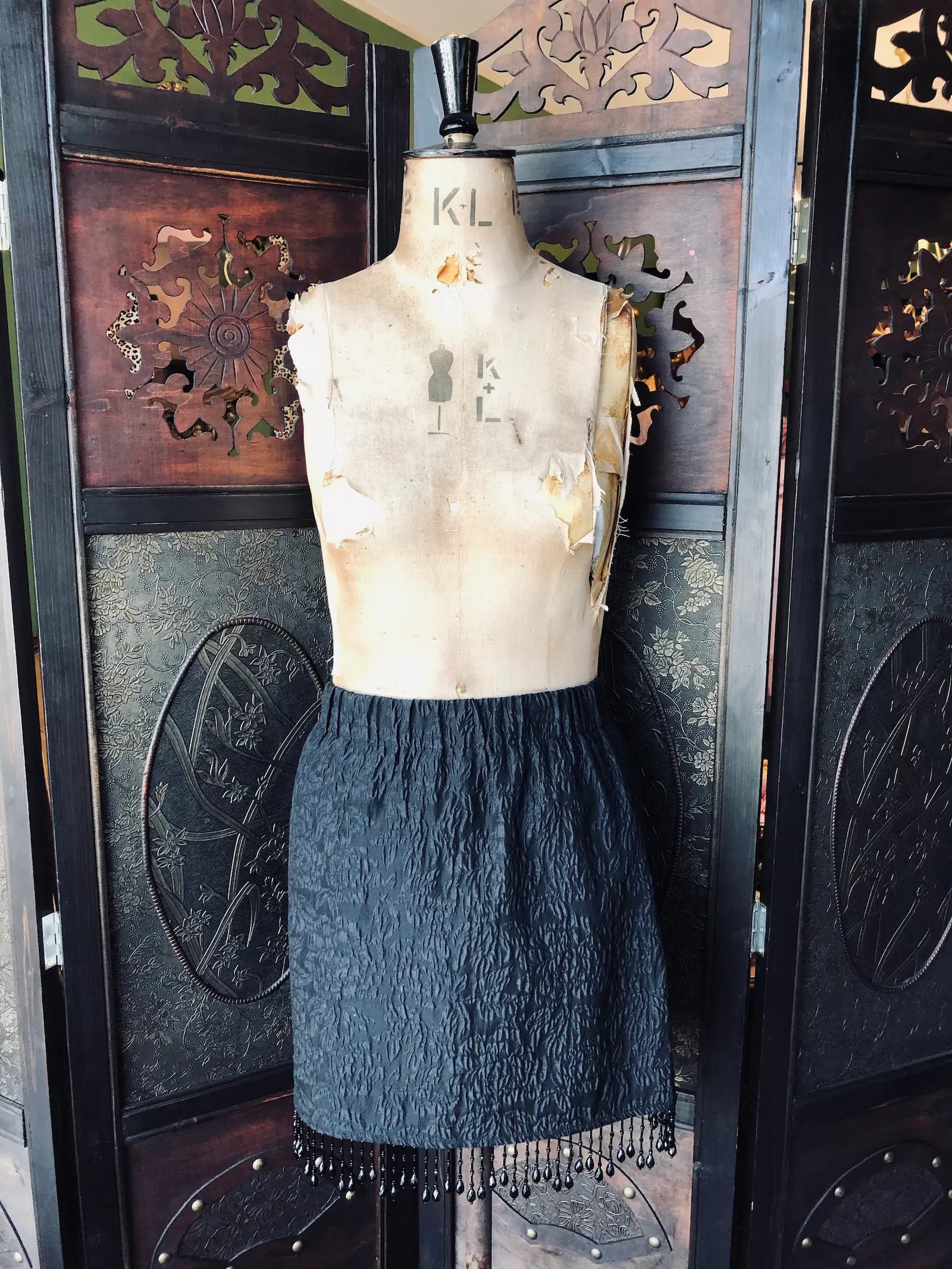 Broochella Mannequin wearing a Ganni black textured skirt with fringe against an ornate wooden background