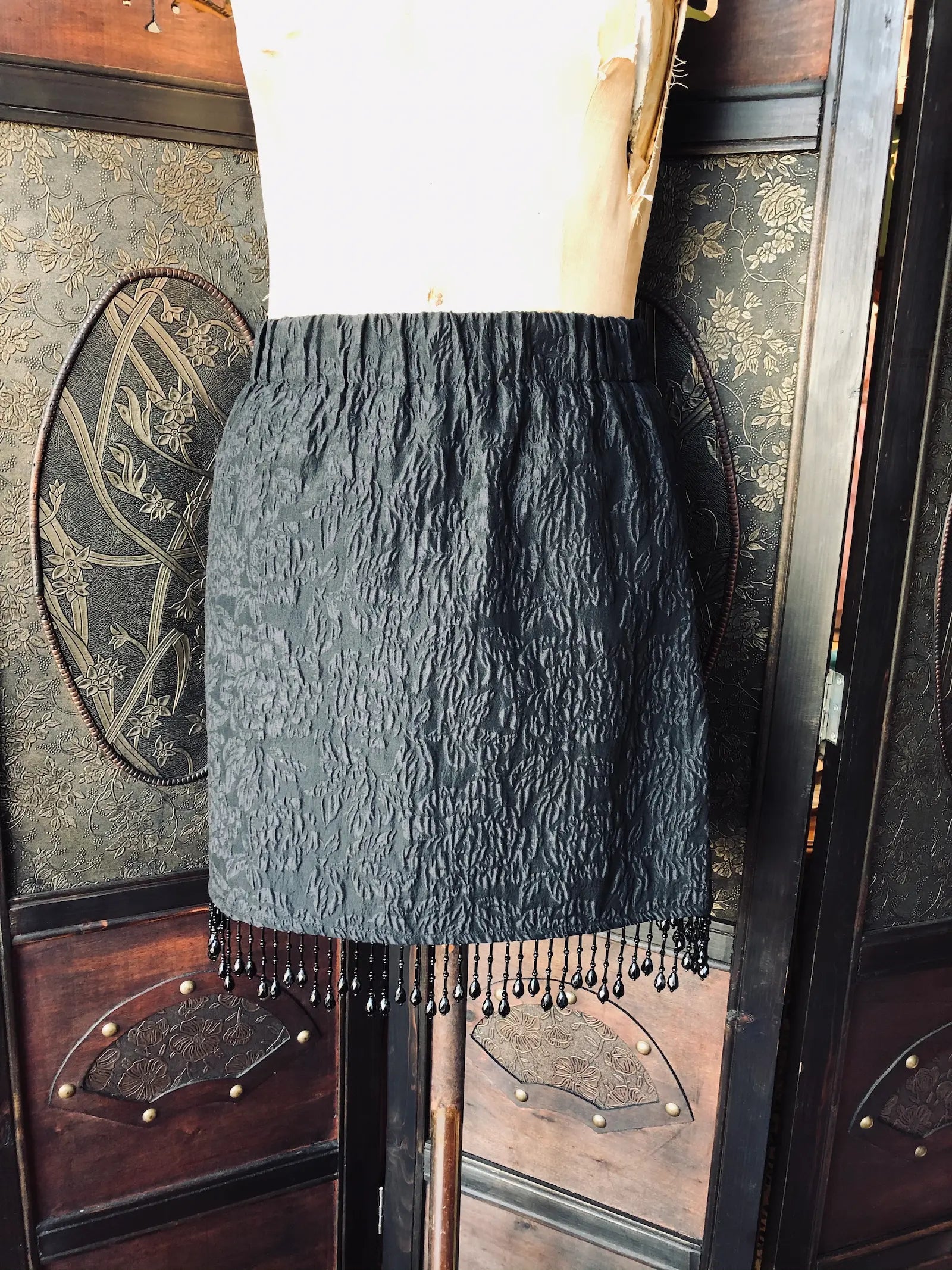 Broochella Mannequin wearing a Ganni black textured skirt with fringe against an ornate wooden background