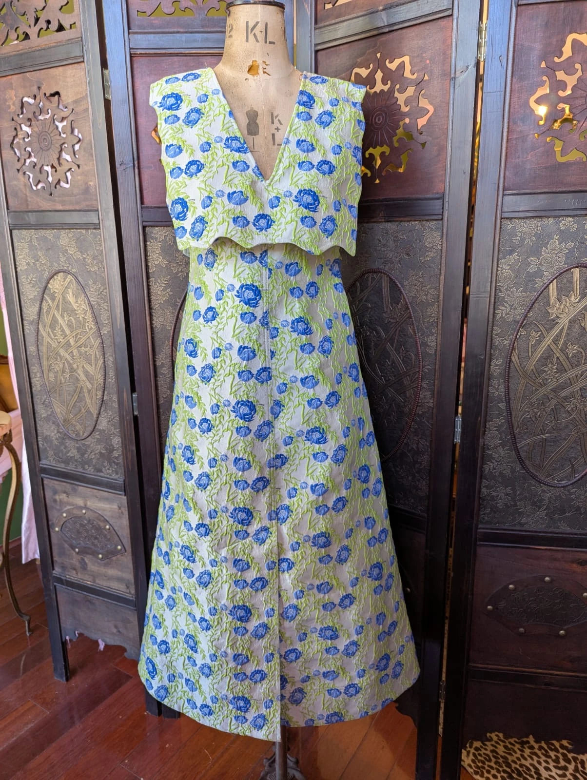 A Broochella Stylist's Closet Ganni sleeveless V-neck dress displayed on a mannequin torso, featuring a blue floral pattern on a pale green textured fabric, set against a dark wooden folding screen.