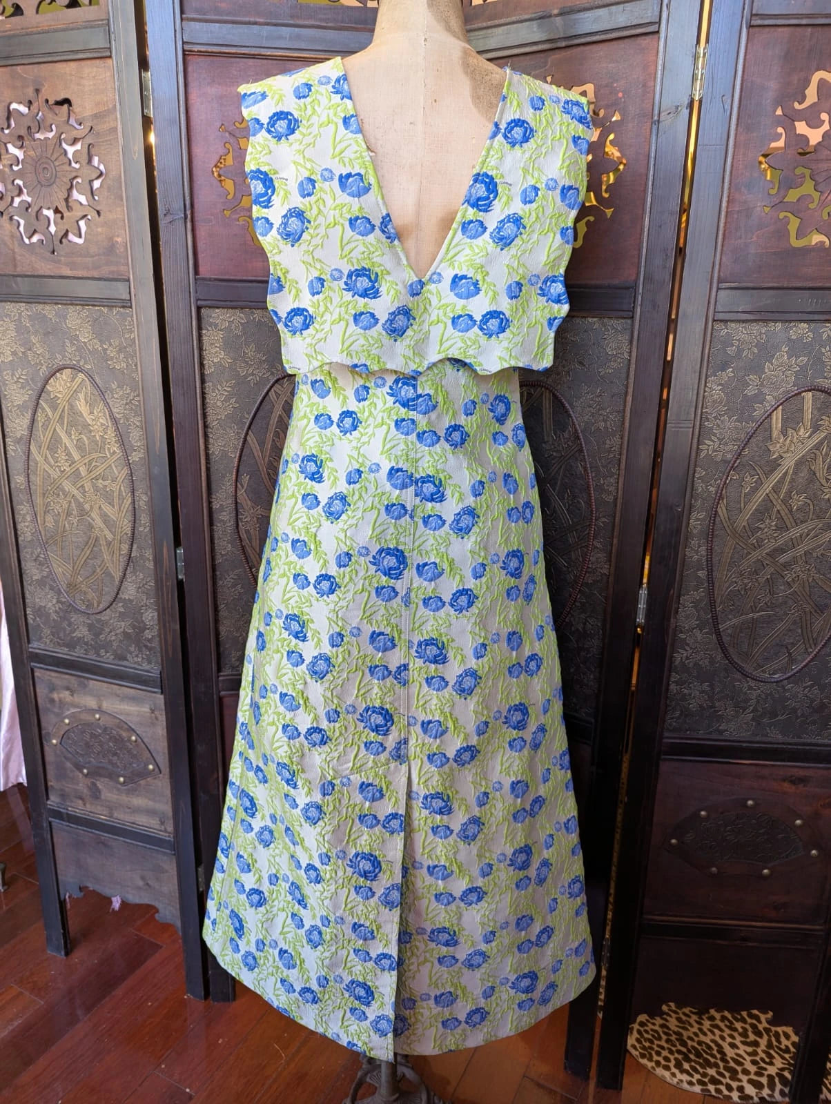 A Broochella Stylist's Closet Ganni sleeveless V-neck dress displayed on a mannequin torso, featuring a blue floral pattern on a pale green textured fabric, set against a dark wooden folding screen.