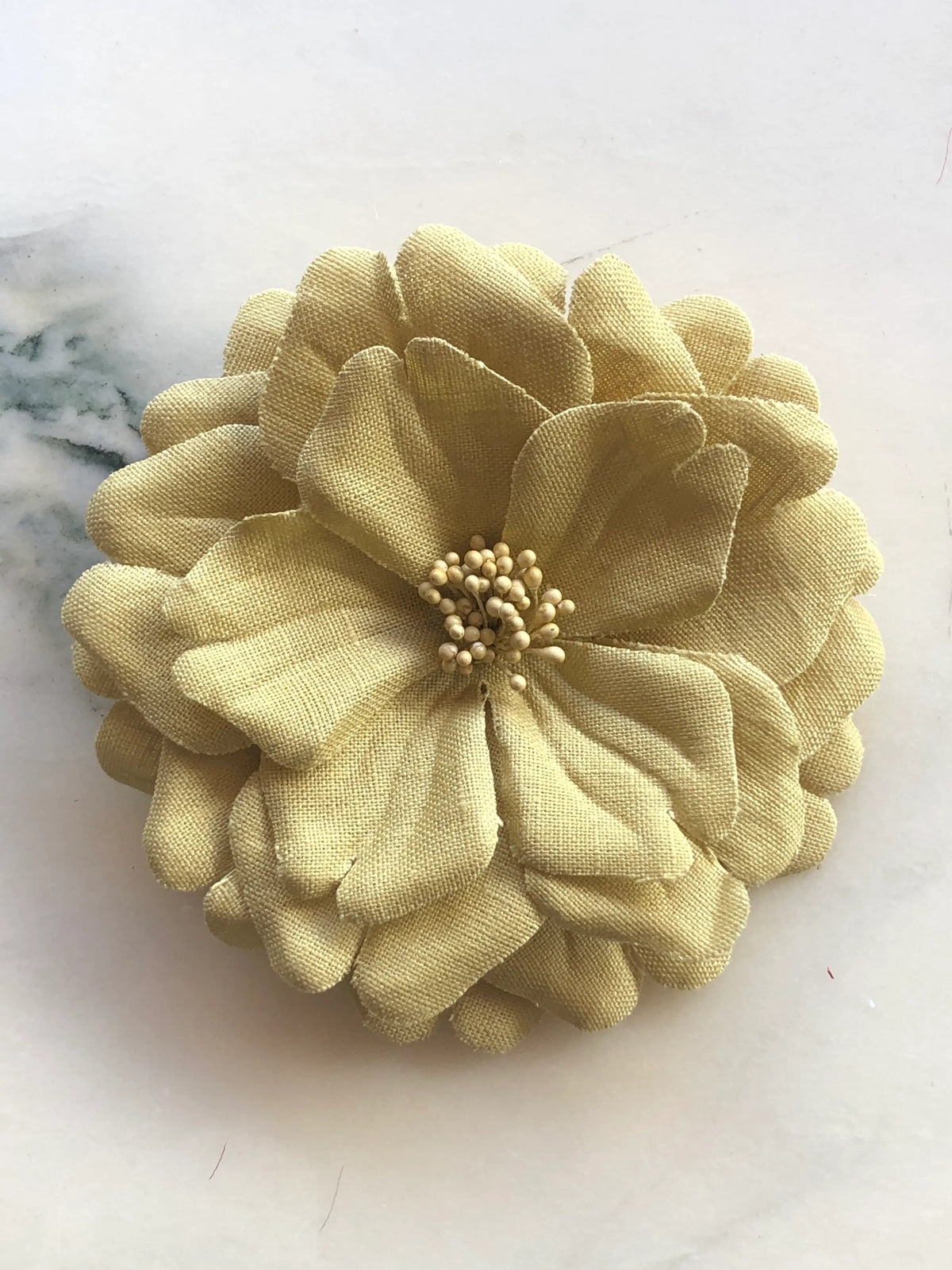 Broochella Garofano Flower Brooch in beige, featuring full layered fabric petals with a textured stamen centre, photographed on a cream marble background.