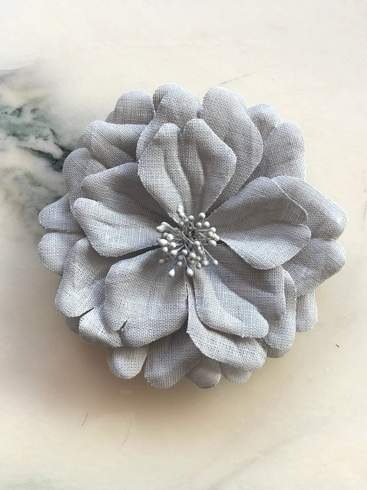 Broochella Garofano Flower Brooch in soft grey, featuring full layered fabric petals with a tonal textured stamen centre, photographed on a cream marble background.