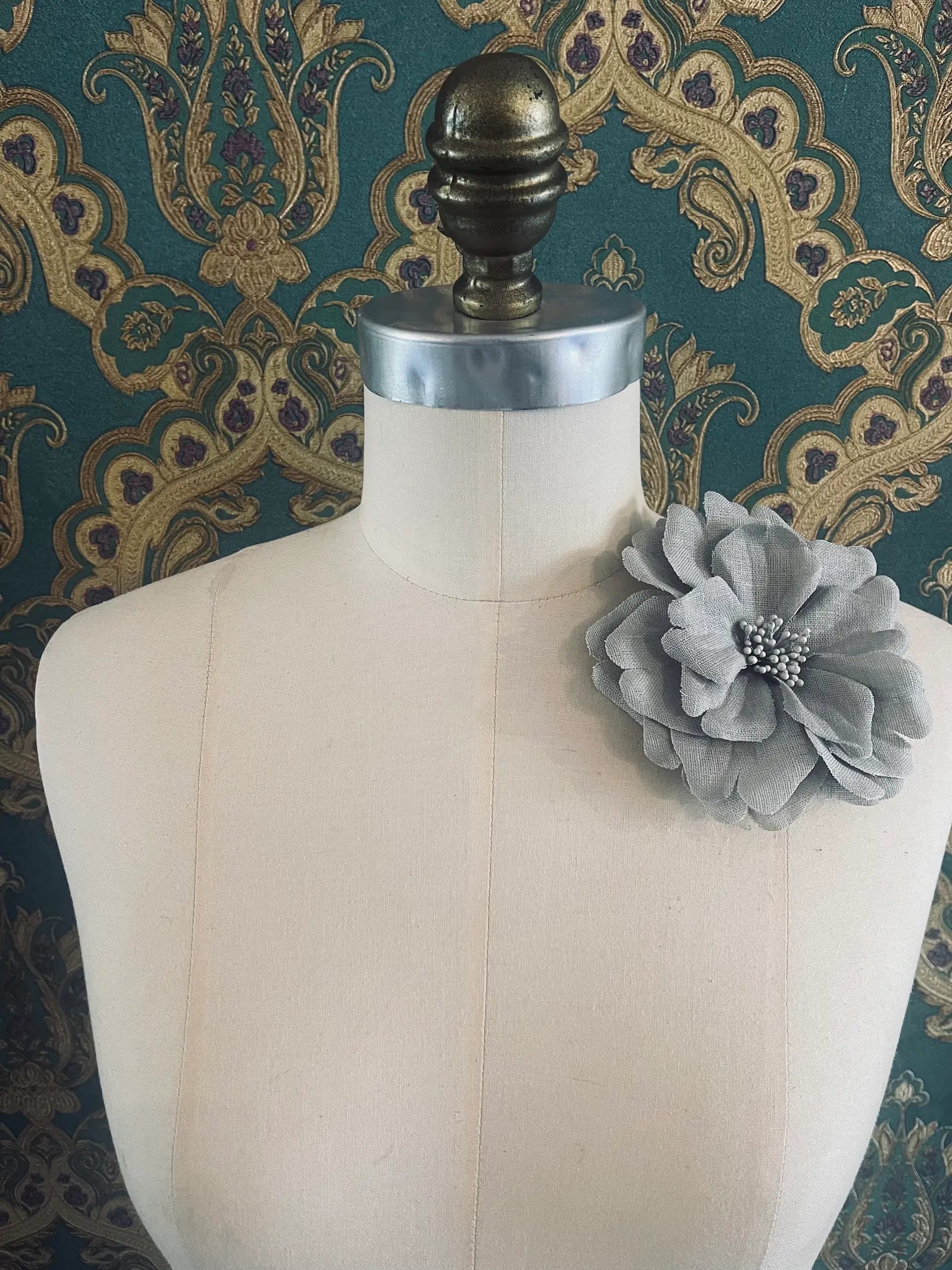 Broochella Garofano handmade fabric flower brooch in grey. Styled on a mannequin. The background is green and gold damask wallpaper