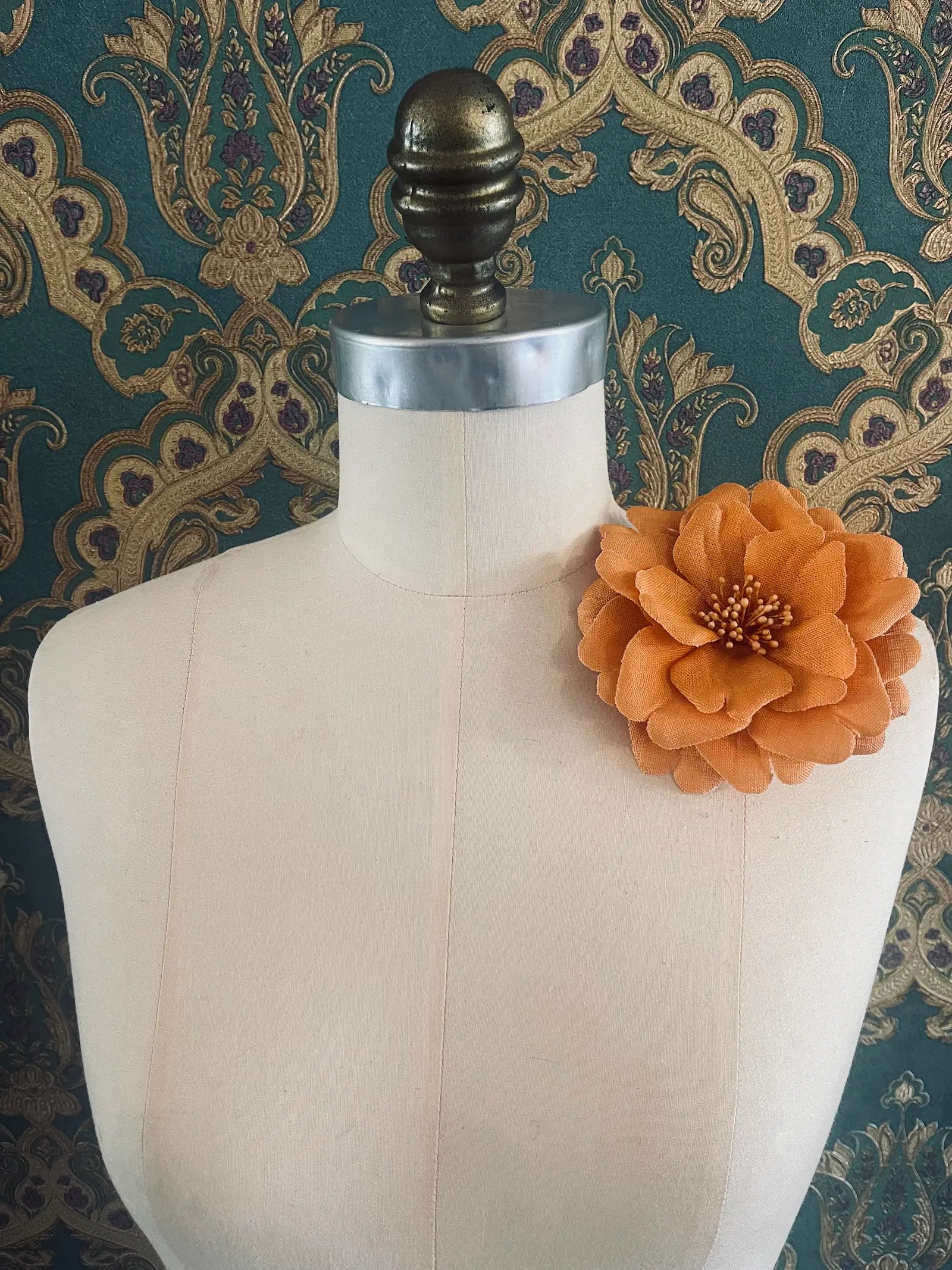 Broochella garofano handmade fabric flower brooch in orange. Styled on a mannequin. The background is green and gold damask wallpaper