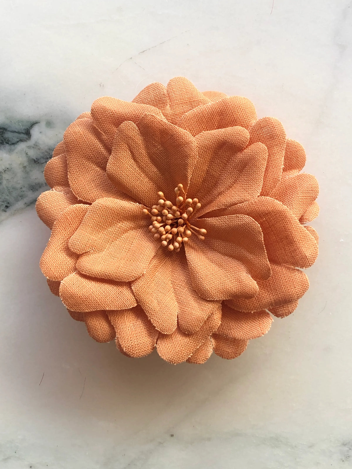 Broochella Garofano Flower Brooch in warm orange, featuring full layered fabric petals with a tonal textured stamen centre, photographed on a cream marble background.