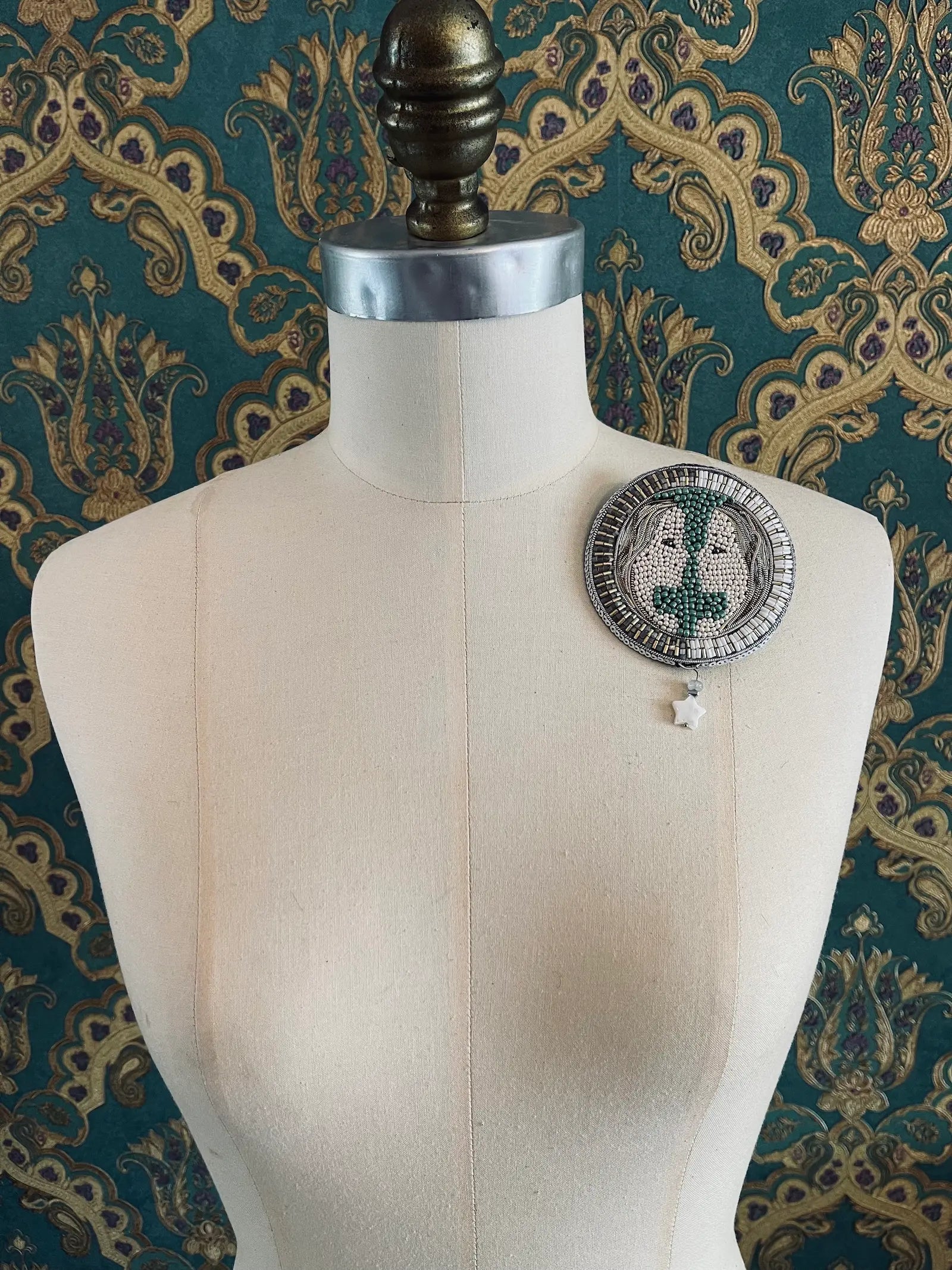 Broochella Gemini hand beaded faces zodiac brooch. Styled on a mannequin. The background is green and gold damask wallpaper