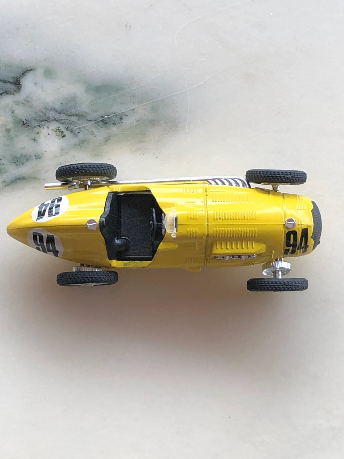 Broochella Giallo Fuoco No.94 Vintage Formula Racing Car Brooch photographed from above, featuring a bright yellow elongated racing car body, black open cockpit, number 94 decals, detailed bonnet vents and four exposed wheels.