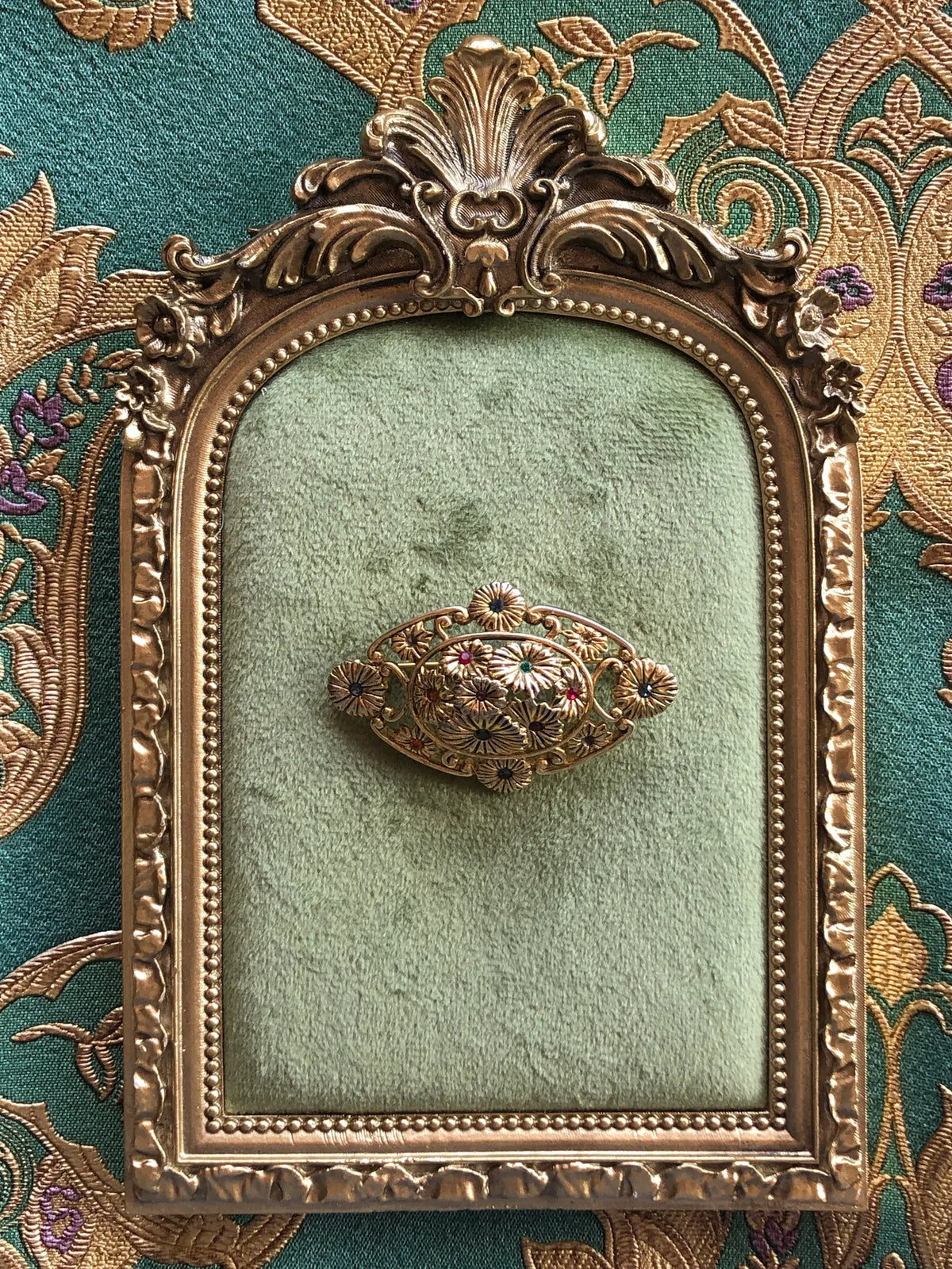 Broochella Giardino Fiorito Vintage Brooch Pin, an ornate gold-tone floral brooch with sculpted flowers and multicoloured crystal centres, displayed on sage green velvet in an antique-style gold frame against green and gold patterned fabric.