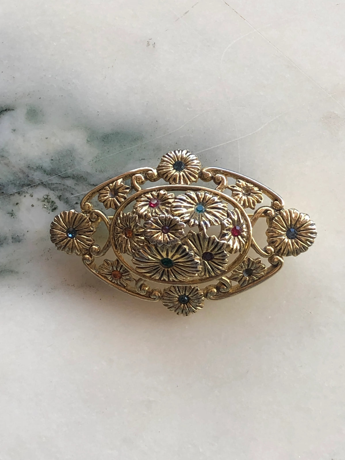 Broochella Giardino Fiorito Vintage Brooch Pin featuring an oval openwork gold-tone design of layered flowers with red, green, blue and amber crystal centres, photographed on white marble with grey-green veining.