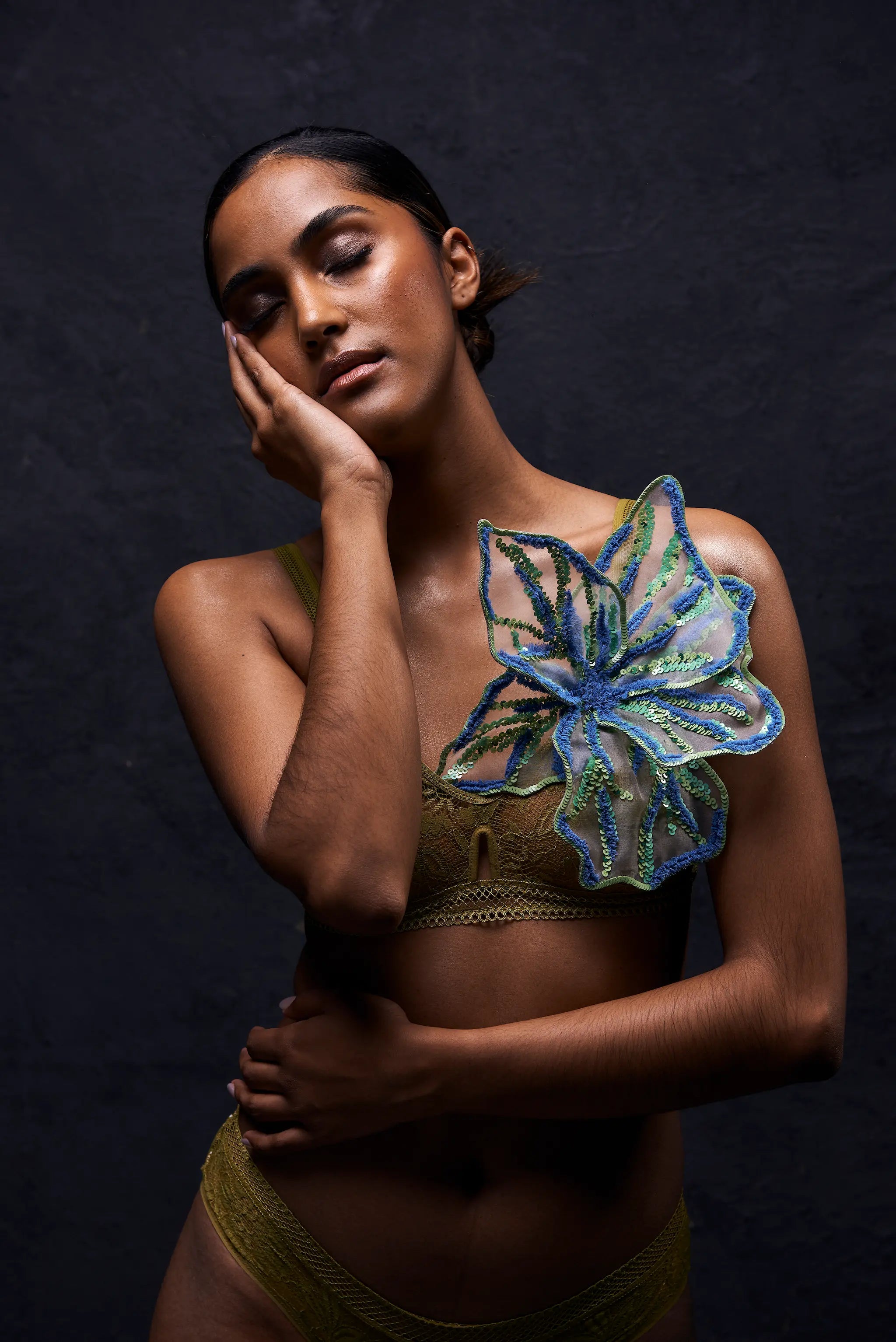 A model wearing a textured olive green bralette with a large, Broochella Giglio di Tigre sculptural blue and green floral brooch on the right shoulder, posed against a dark, textured studio background.