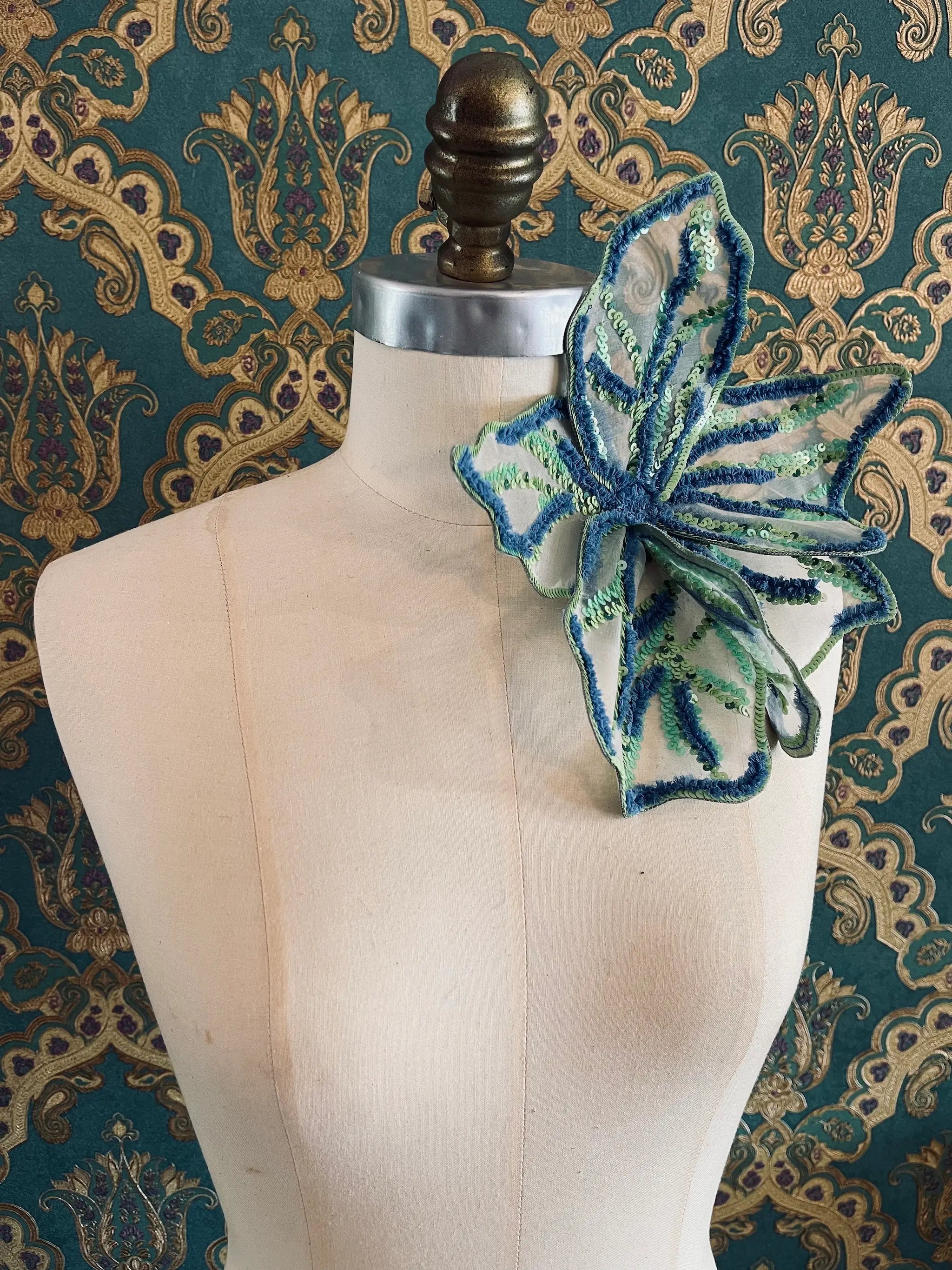 Broochella Giglio di Tire hand beaded flower brooch in blue and green. Pictured on a mannequin. The background is green and gold damask wallpaper