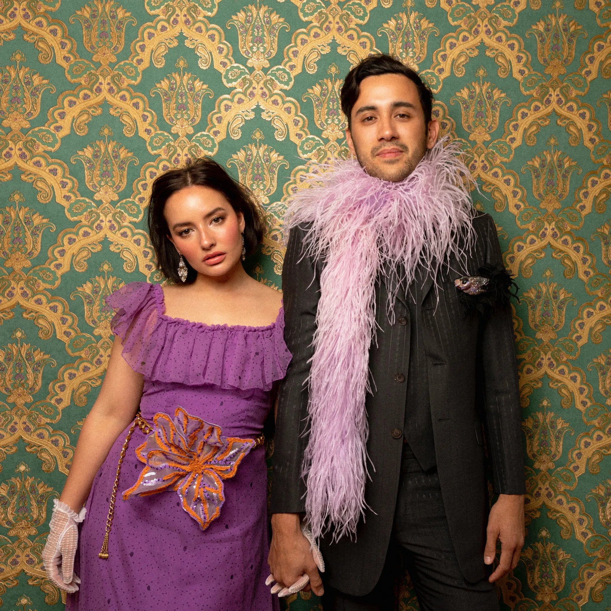A male and female model in a suit and a purple dress. The lady is wearing the Broochella Giglio di tigre brooch in purple and orange. The man is wearing a lilac feather boa. The background is green and gold damask wallpaper