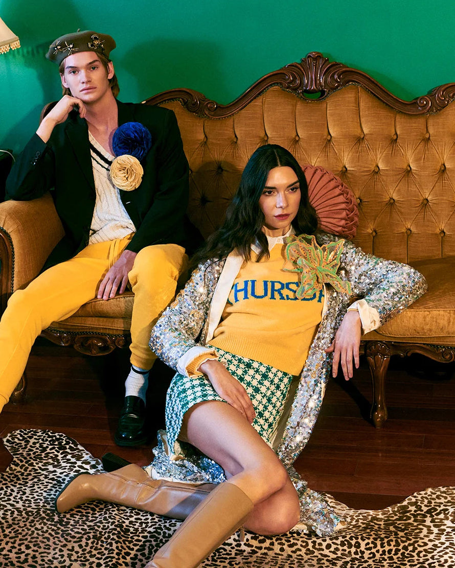 A fashion editorial photo featuring a male and female model in retro 1960s-inspired outfits. The man wears a black blazer, white shirt, yellow trousers, and a beret. She is wearing the Broochella large Giglio di Tigre hand beaded brooch in green. 
