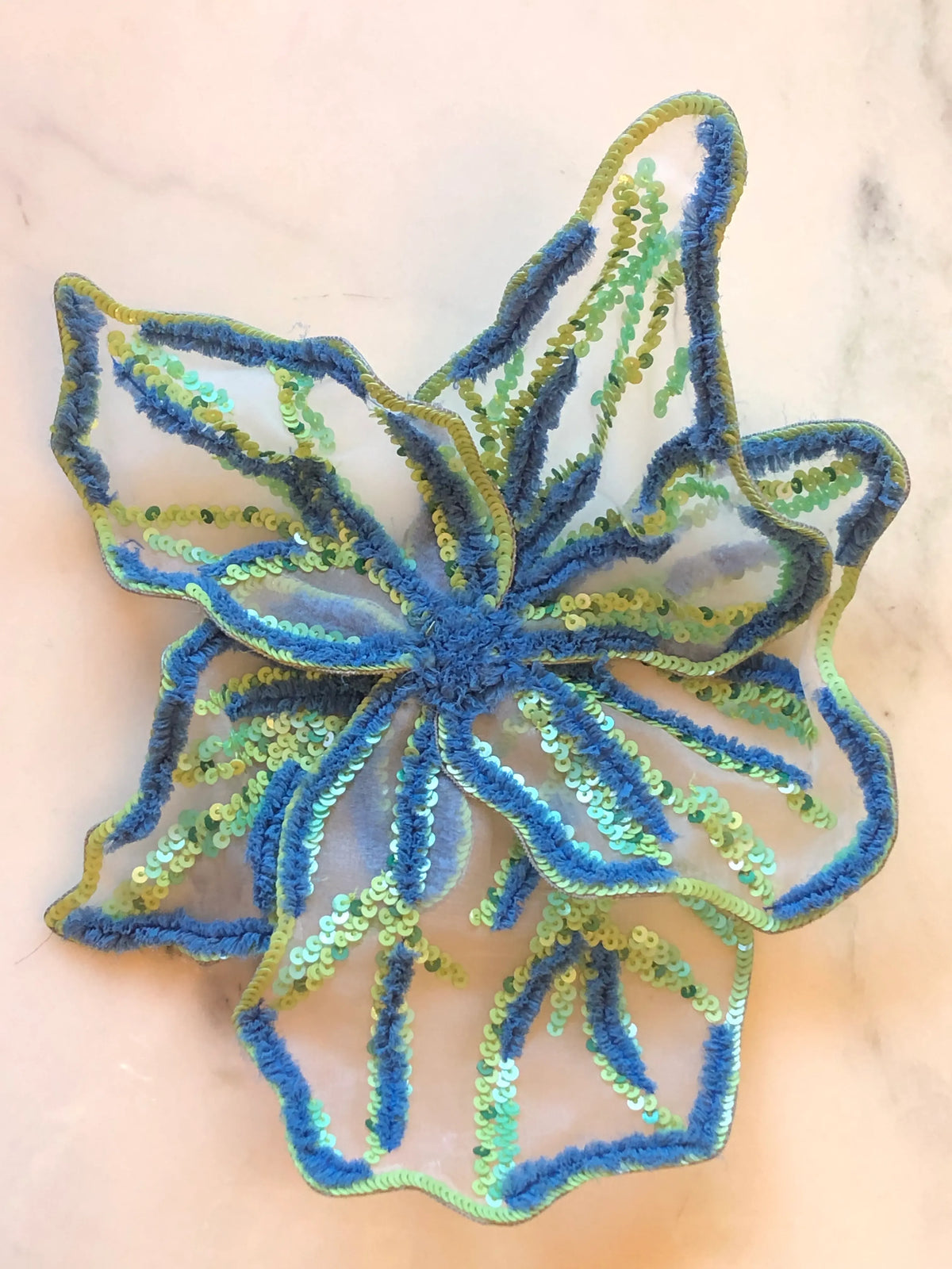Broochella Giglio di Tigre Flower Brooch Pin - Large in blue, featuring sculptural sheer petals embellished with blue, chartreuse and aqua sequins and textured embroidery on a cream marble background.