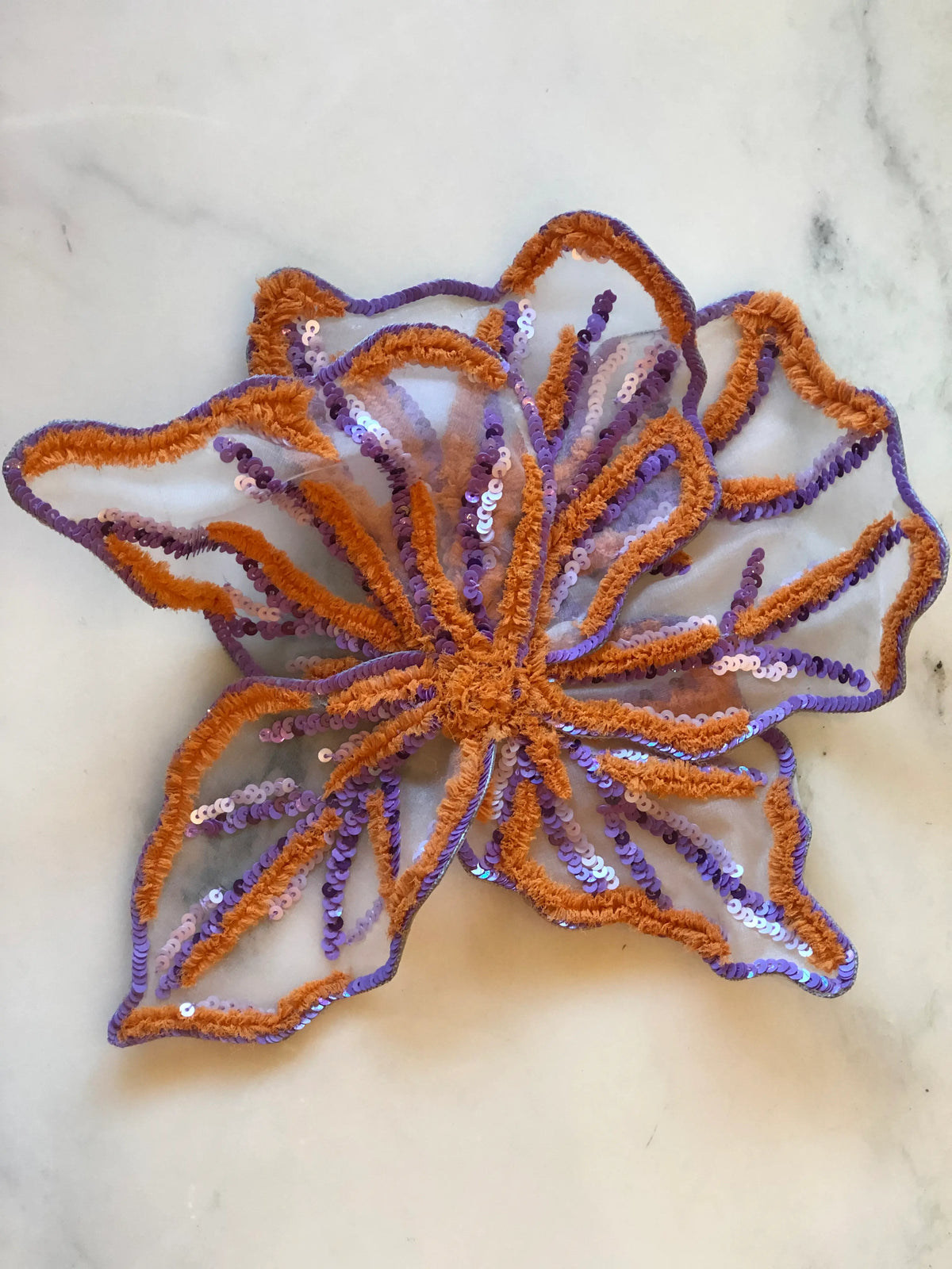 Broochella Giglio di Tigre Flower Brooch Pin - Large in purple, featuring sculptural sheer petals embellished with purple and lilac sequins and contrasting burnt orange textured embroidery on a cream marble background.