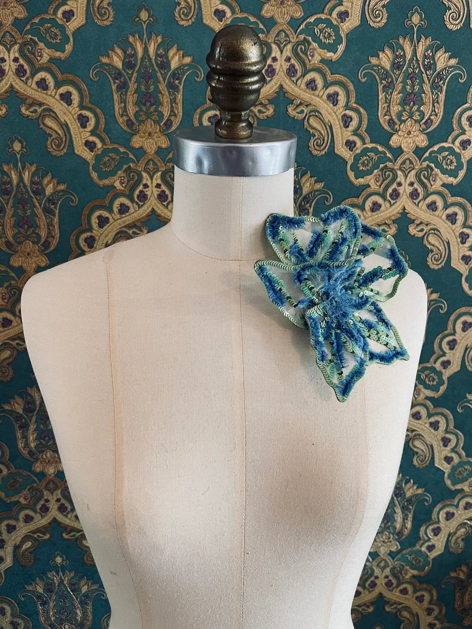 Broochella giglio di tigre small hand beaded, sequinned, flower brooch in blue and green. It is pinned to a mannequin. The background is green and gold damask wallpaper