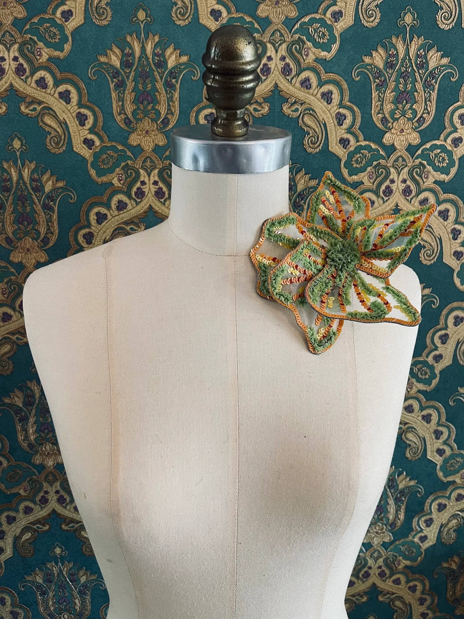 Broochella giglio di tigre small hand beaded, sequinned, flower brooch in green and orange. It is pinned to a mannequin. The background is green and gold damask wallpaper