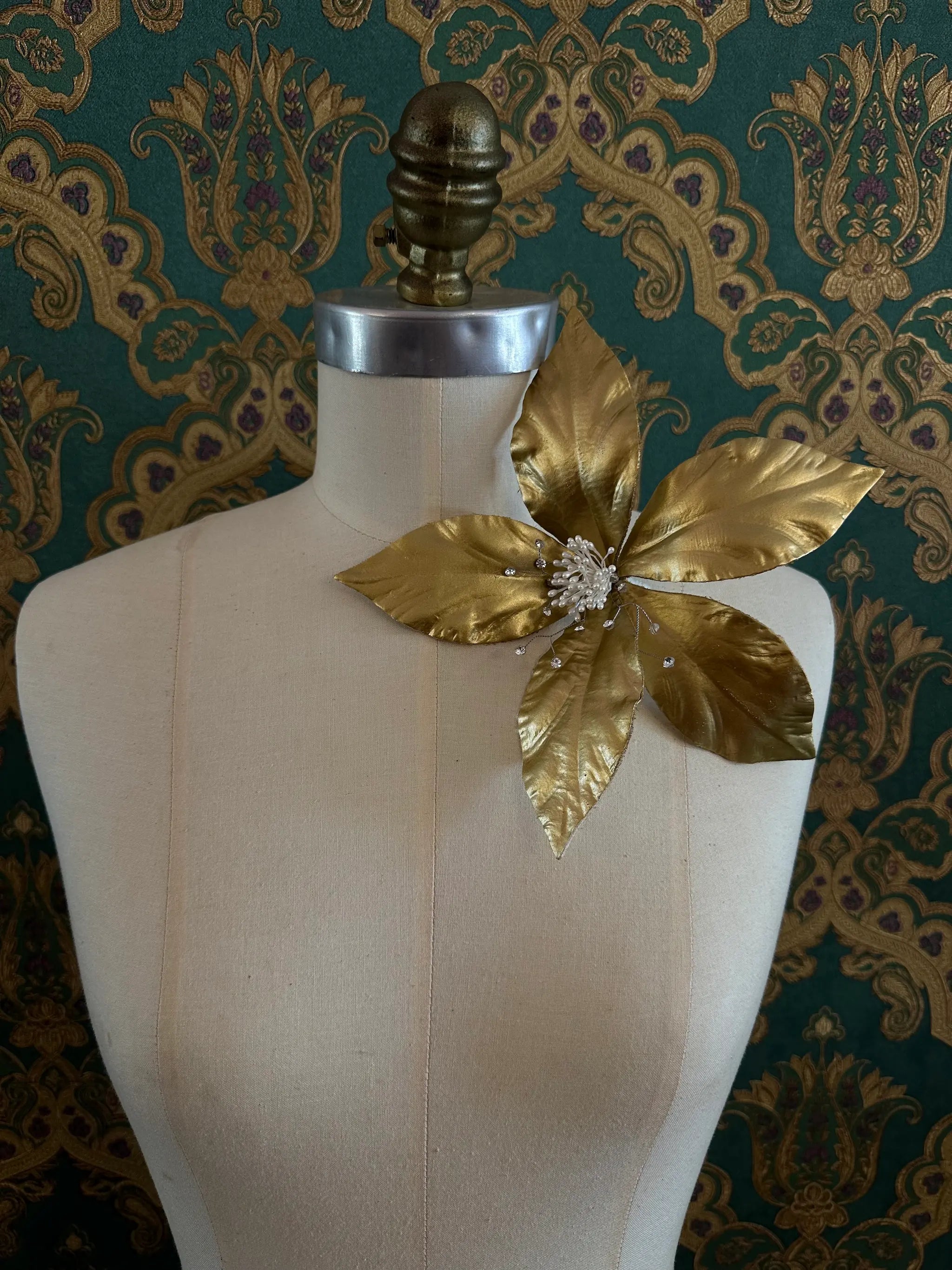 Broochella giglio handmade large patent leather flower in gold. Styled on a mannequin. The background is green and gold damask wallpaper