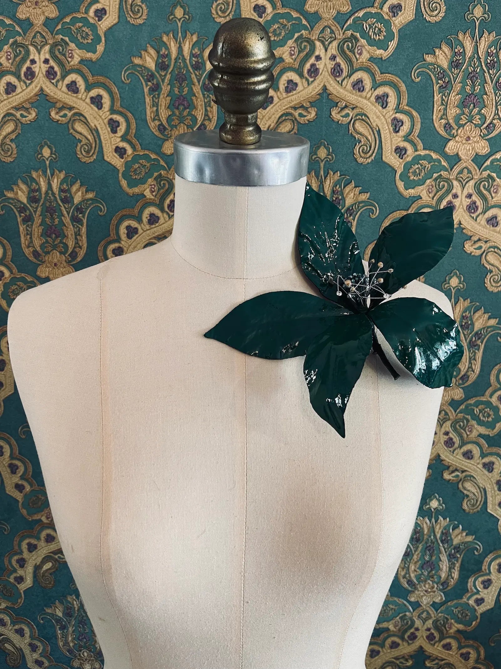 Broochella giglio handmade large patent leather flower in green. Styled on a mannequin. The background is green and gold damask wallpaper