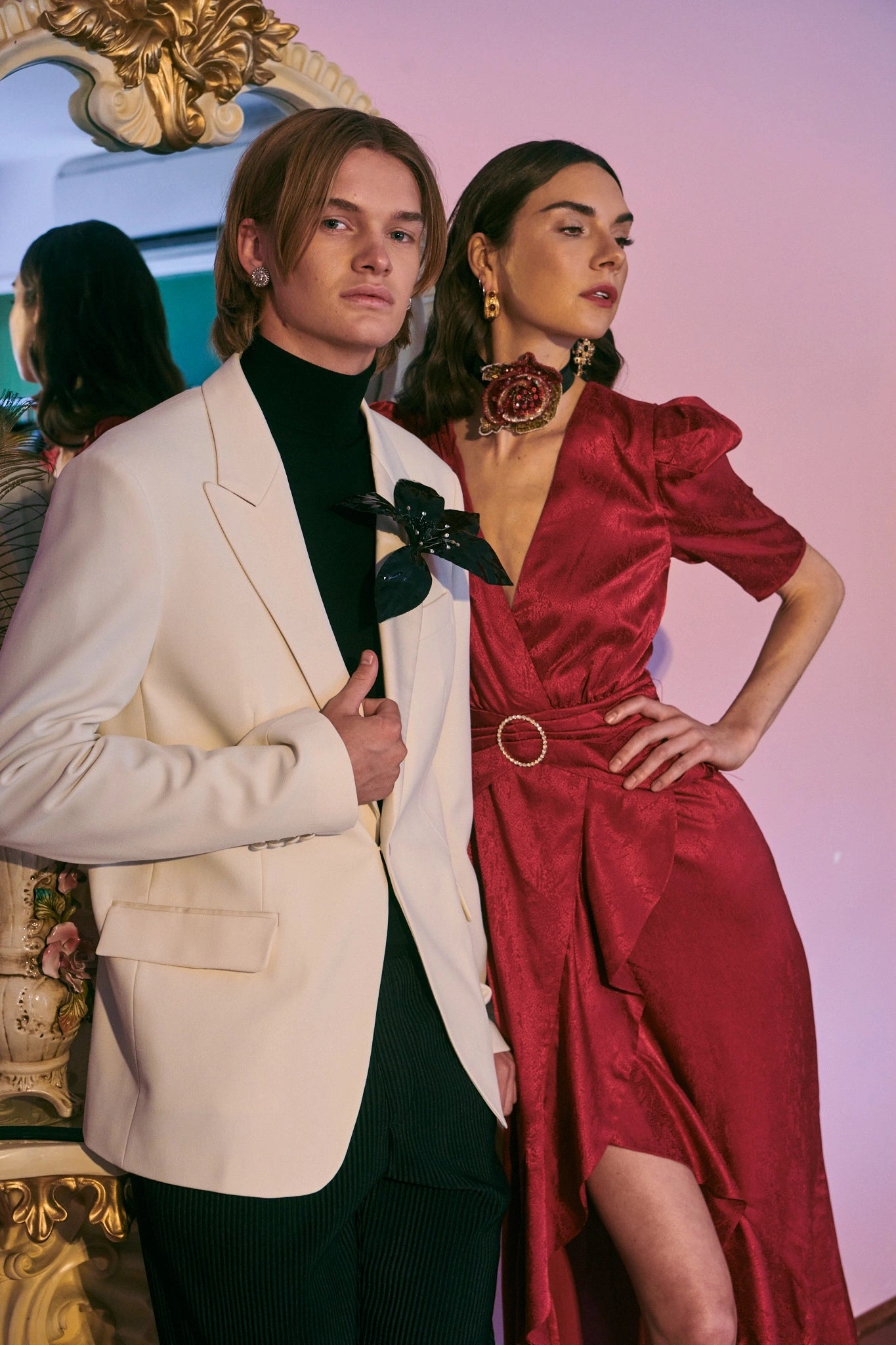 Two models in formal attire standing in front of a decorative mirror. The man is in a white suit jacket. He is wearing the Broochella giglio flower brooch. The lade i in a red satin dress. She has dark hair.