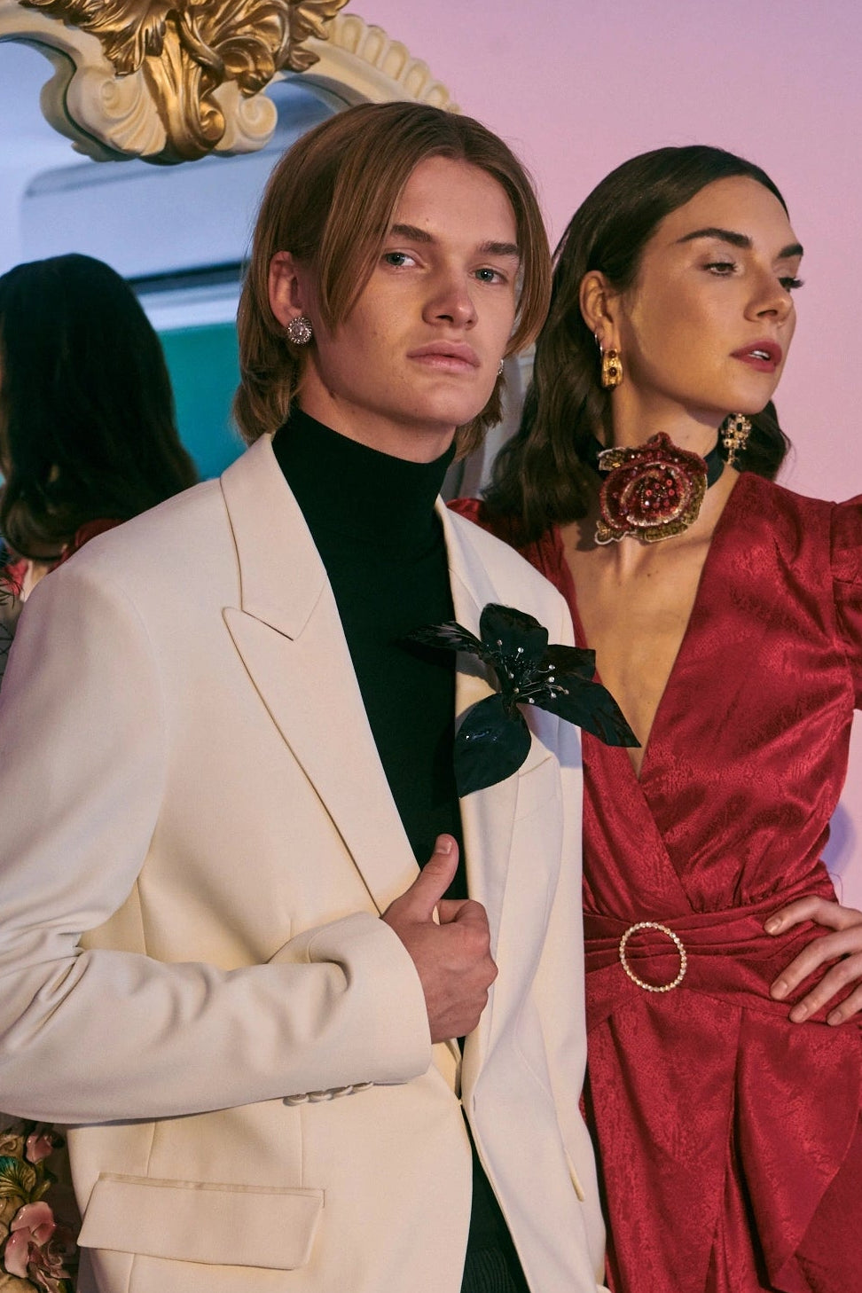 Two models in formal attire standing in front of a decorative mirror. The man is in a white suit jacket. He is wearing the Broochella giglio flower brooch. The lade i in a red satin dress. She has dark hair.